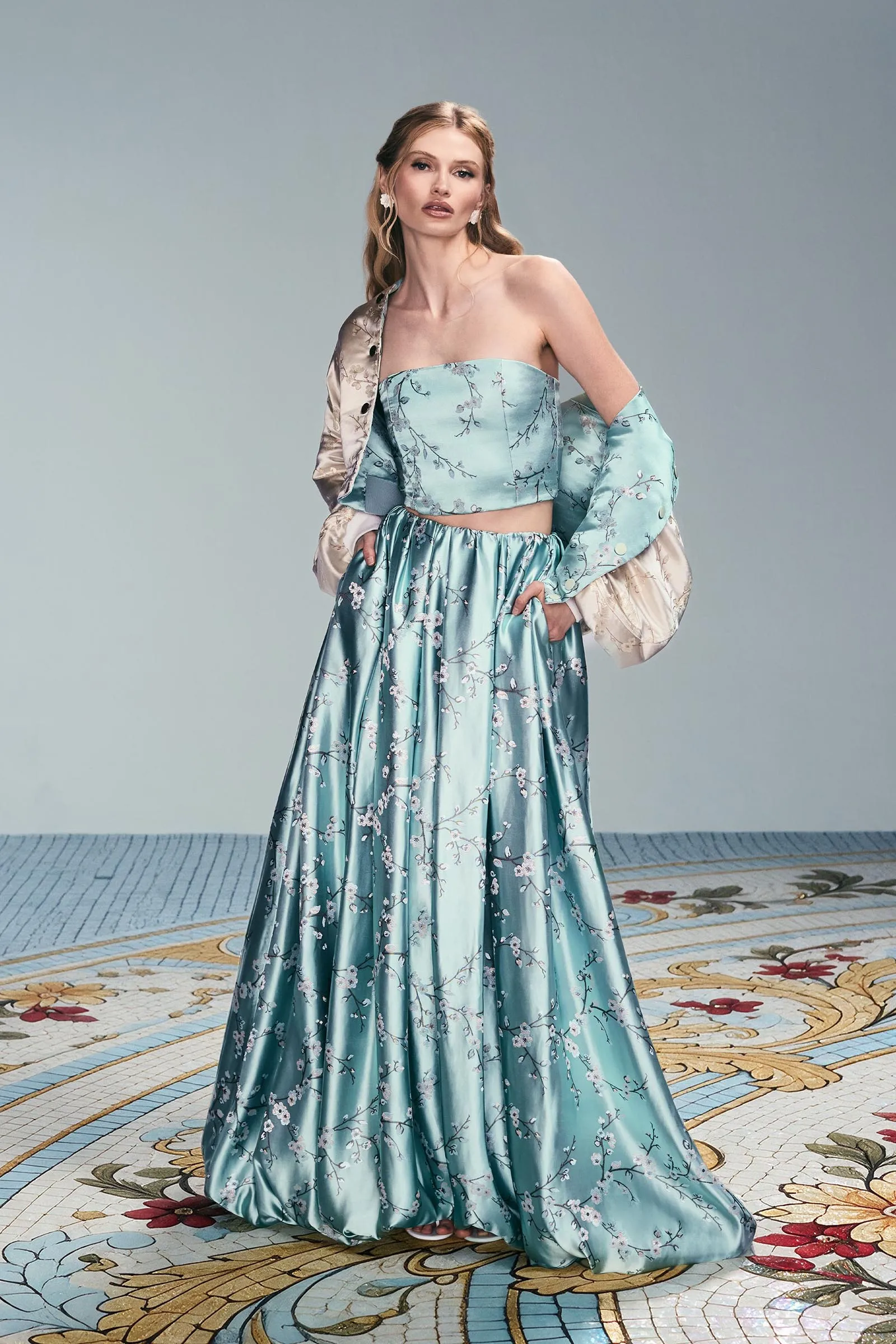 Model wearing a teal floral strapless gown with a matching satin jacket, standing with hands on hips in a studio setting.