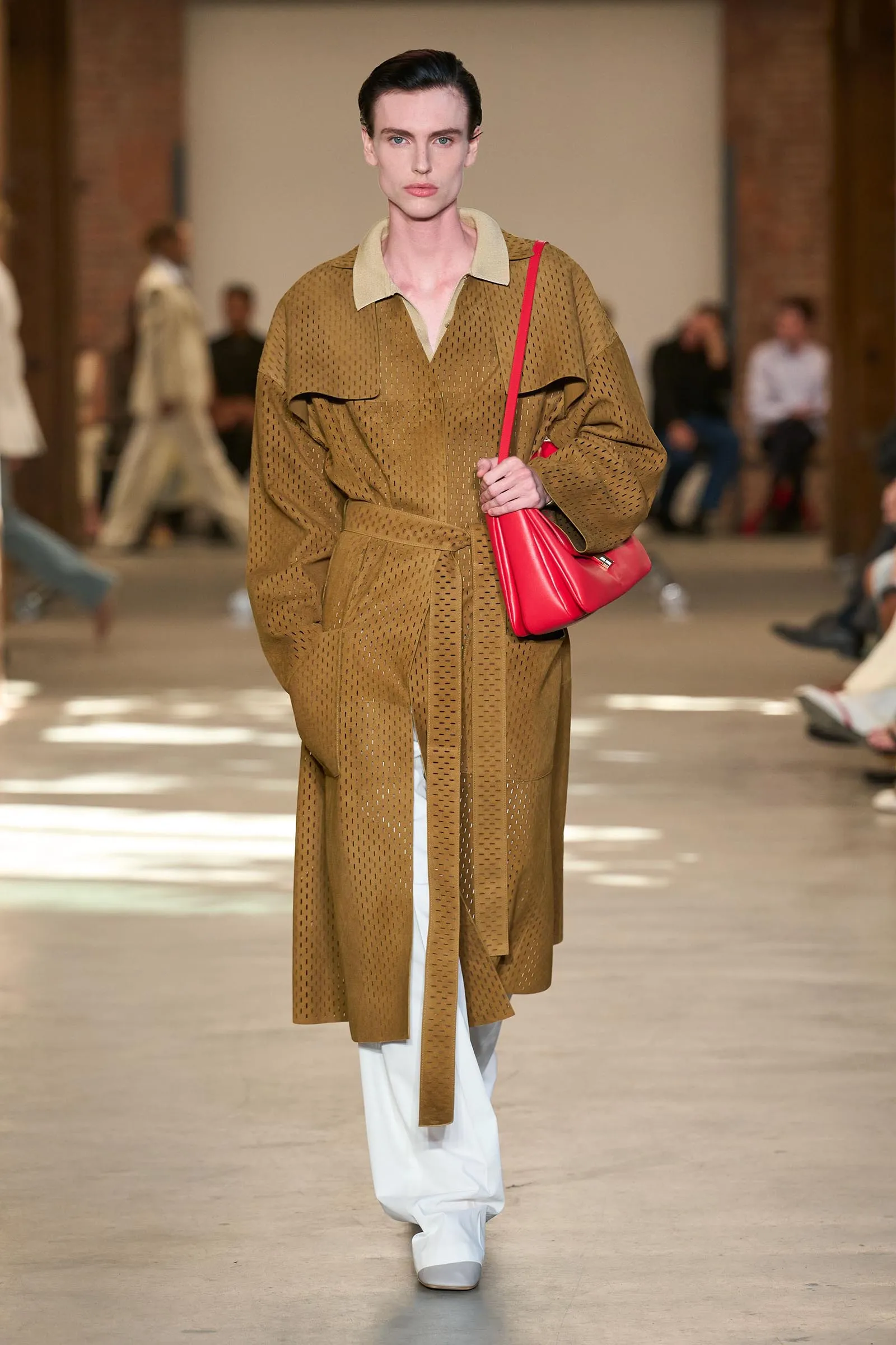 Model on a runway wearing a tan perforated coat with a beige collar, belted at the waist, paired with white pants and a red shoulder bag.