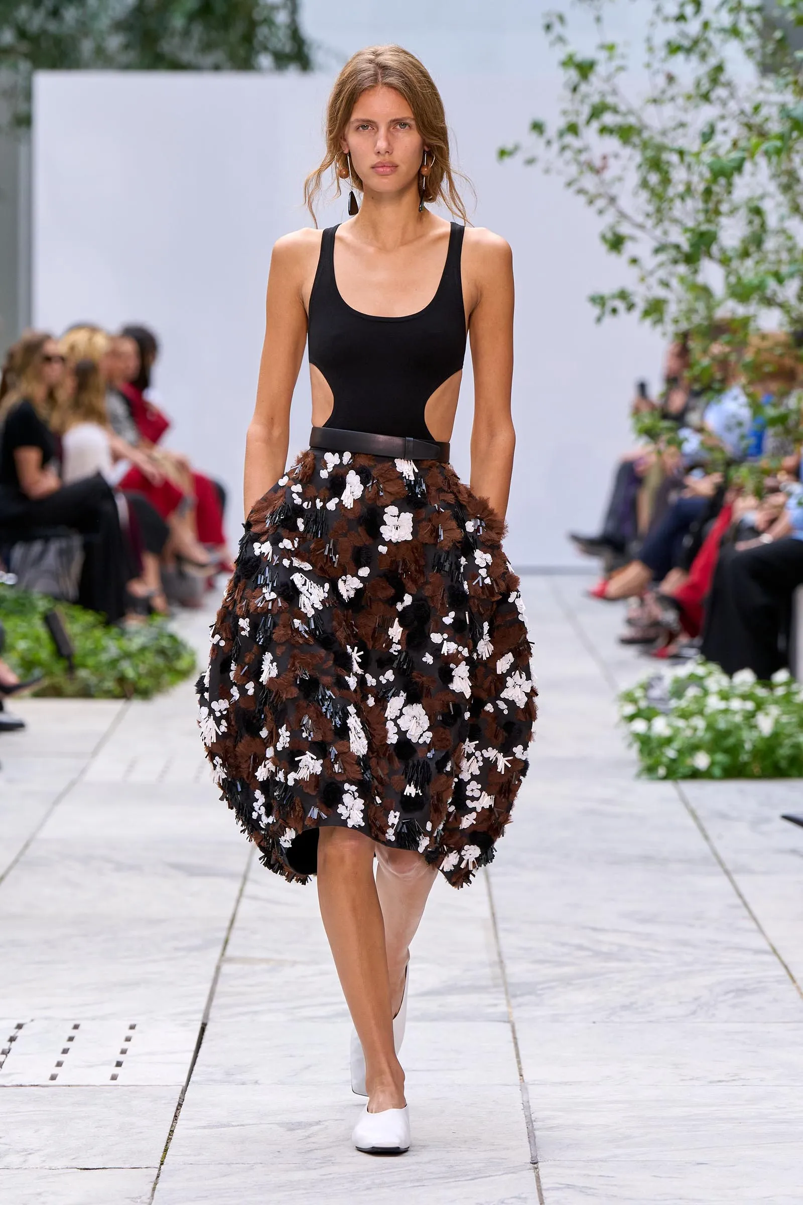 Model walks a daytime runway in a black sleeveless top with side cutouts and a brown, white-flowered feathered skirt, white shoes.