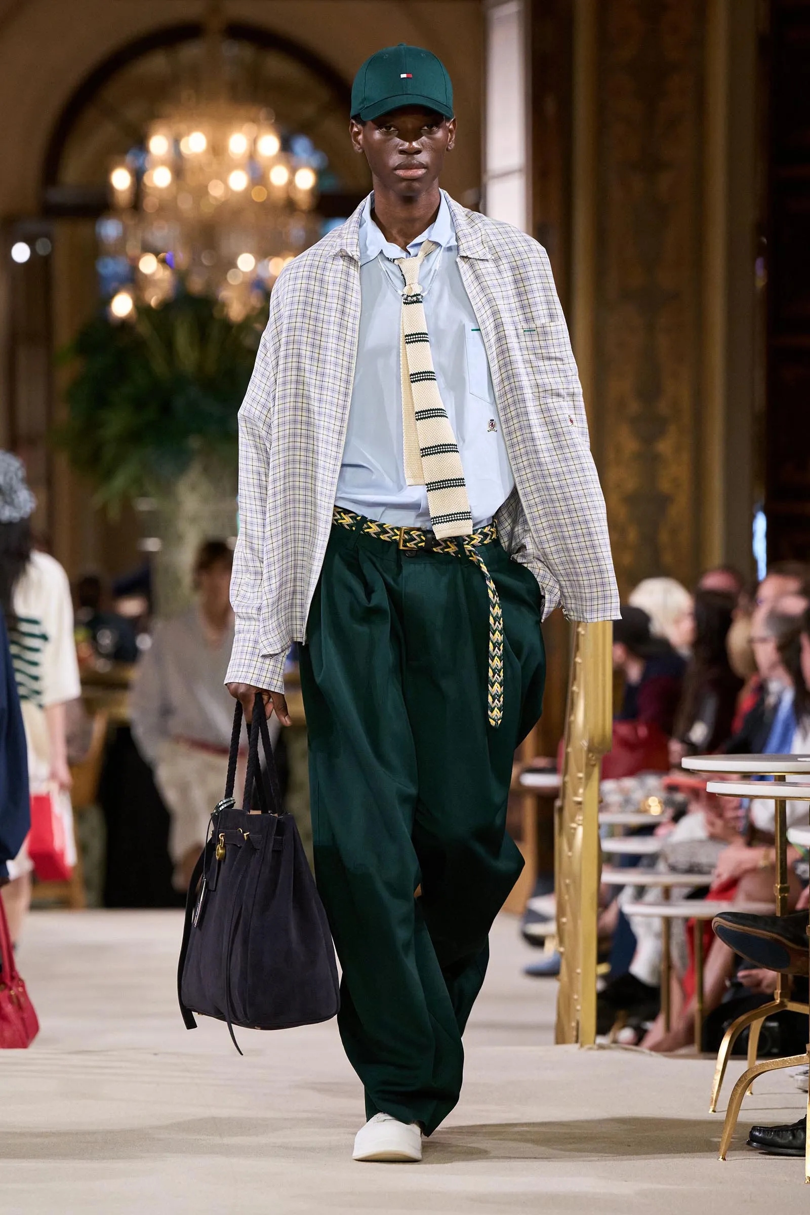 Model walking a fashion runway in a light blue shirt, striped beige tie, plaid overcoat, dark green trousers, white sneakers, and a teal cap with a black bag in hand