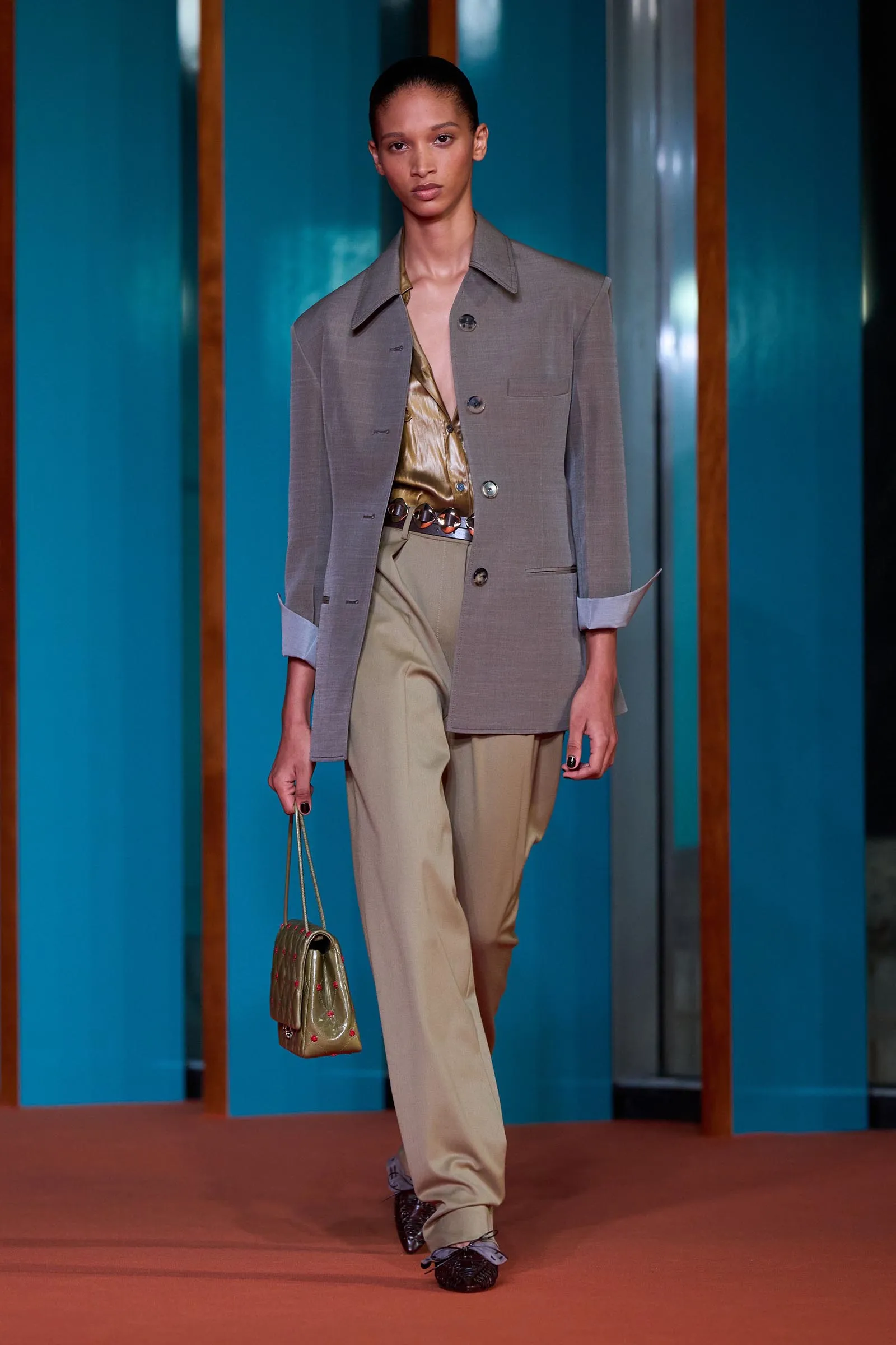 Model walking a runway in a gray blazer, gold top, and beige trousers, carrying a small gold handbag against teal panels.