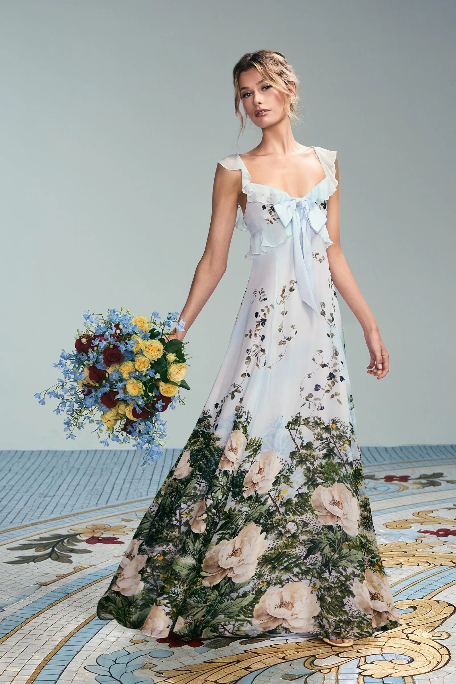 Woman in a light blue floral maxi dress holds a large bouquet of yellow, red, and blue flowers.