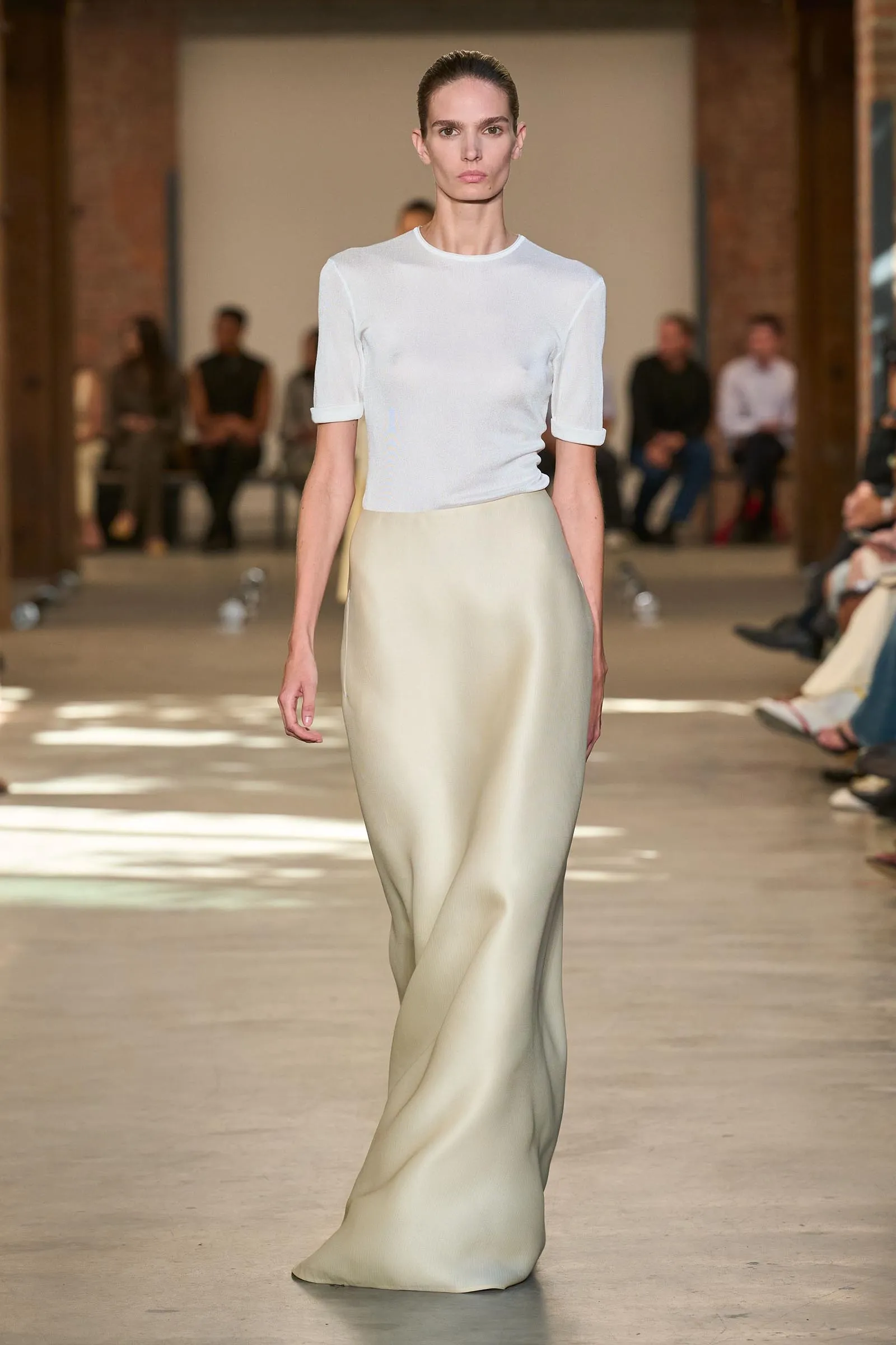 Model walks a fashion runway wearing a white short-sleeve top tucked into a cream satin skirt, audience blurred in the background.