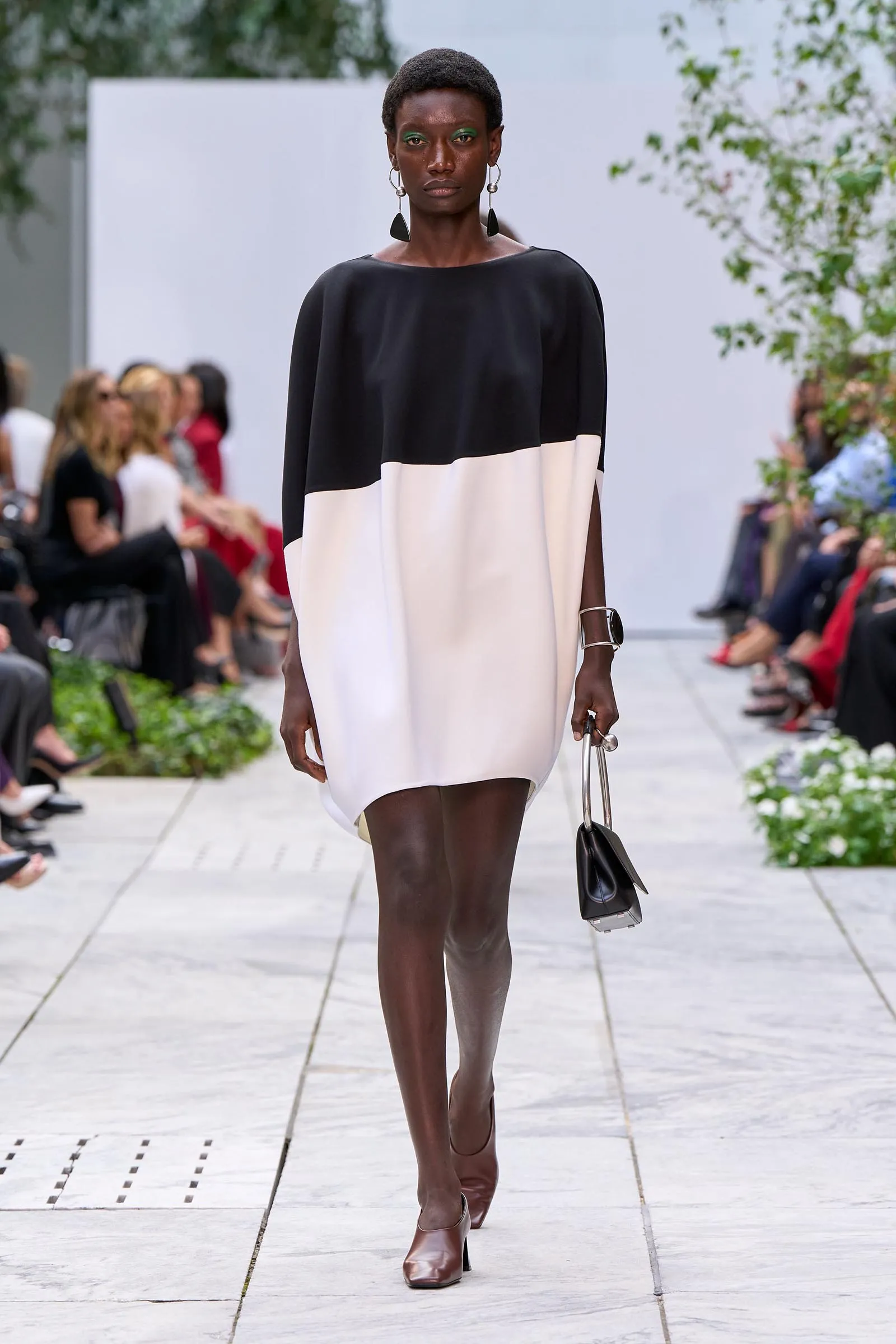 Model walking a runway in a black-and-cream color-block dress, brown ankle boots, bold dangling earrings, and a small black handbag.