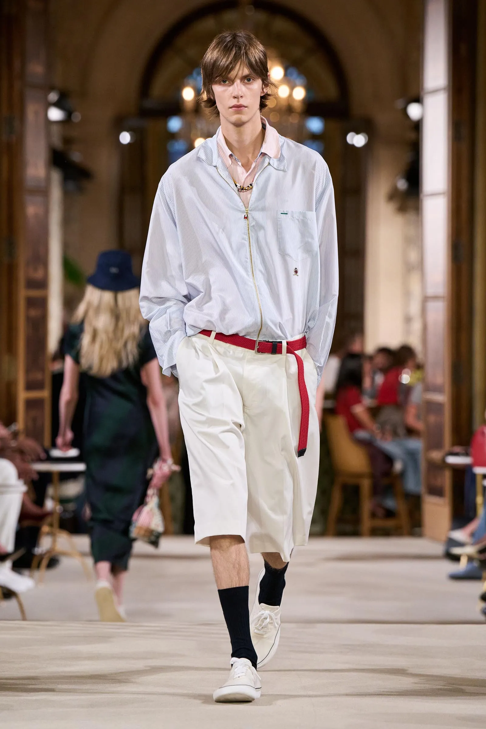Male model walking a runway in a light blue striped zip-up jacket, white shorts, and a red belt, with white sneakers in a fashion show setting.