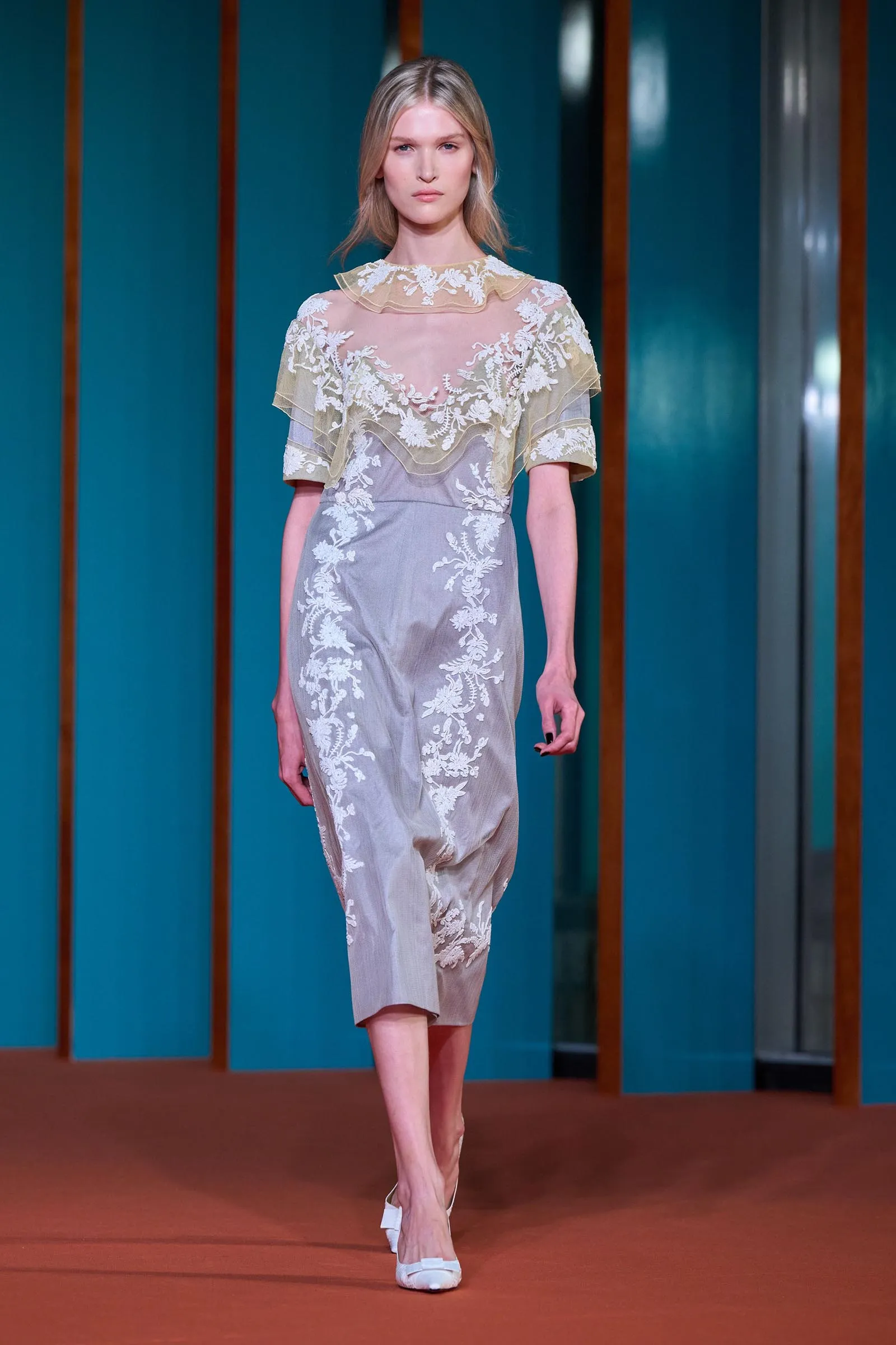 Model walking a runway in a pale sheer embroidered dress with white floral lace appliqués.