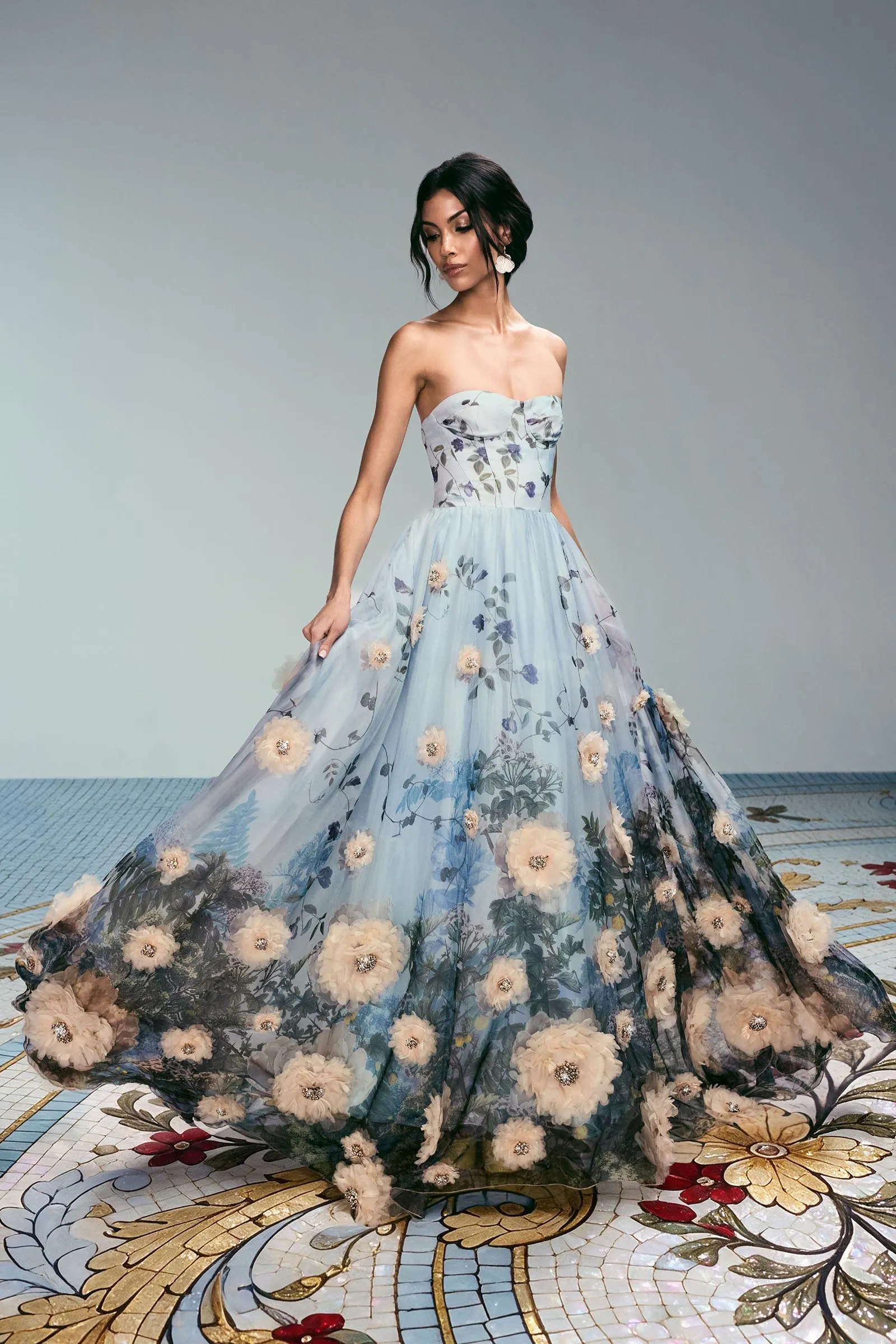 Woman in a strapless light-blue floral ball gown, posing against a pale gradient studio backdrop.