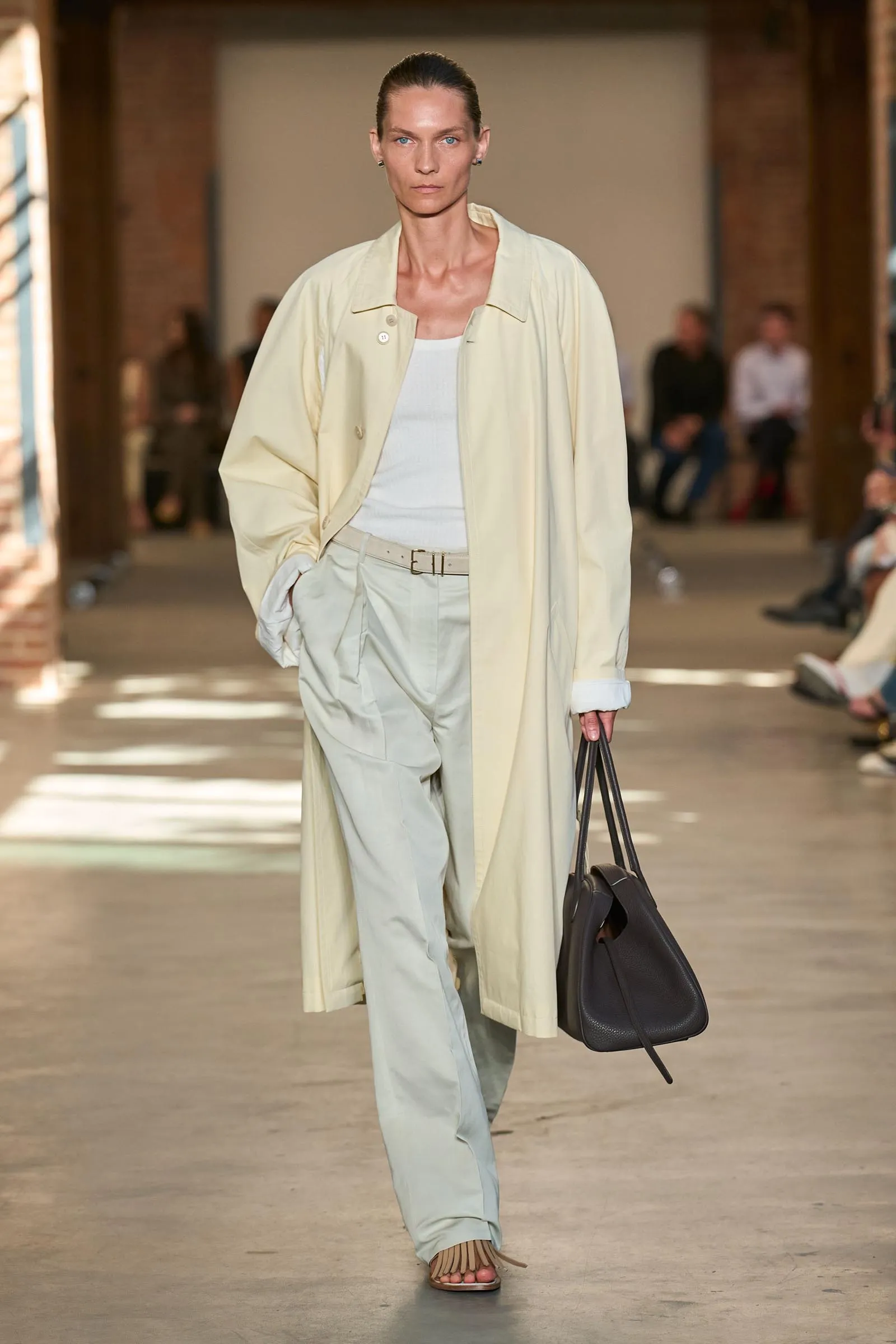 Model walks a fashion runway in a pale beige trench coat over a white top and light pants, carrying a black handbag.