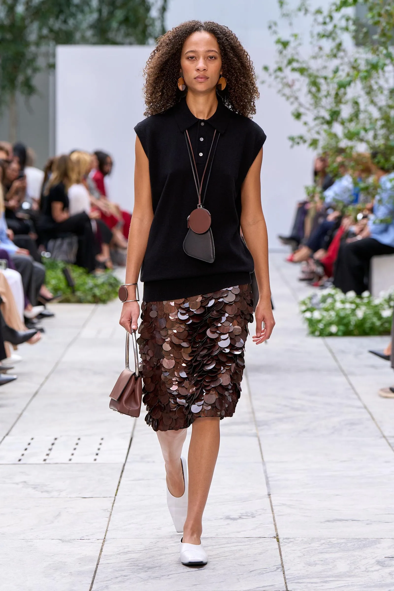 Model walking runway in a black sleeveless top and a brown sequined skirt, carrying a brown handbag and wearing circular brown earrings.