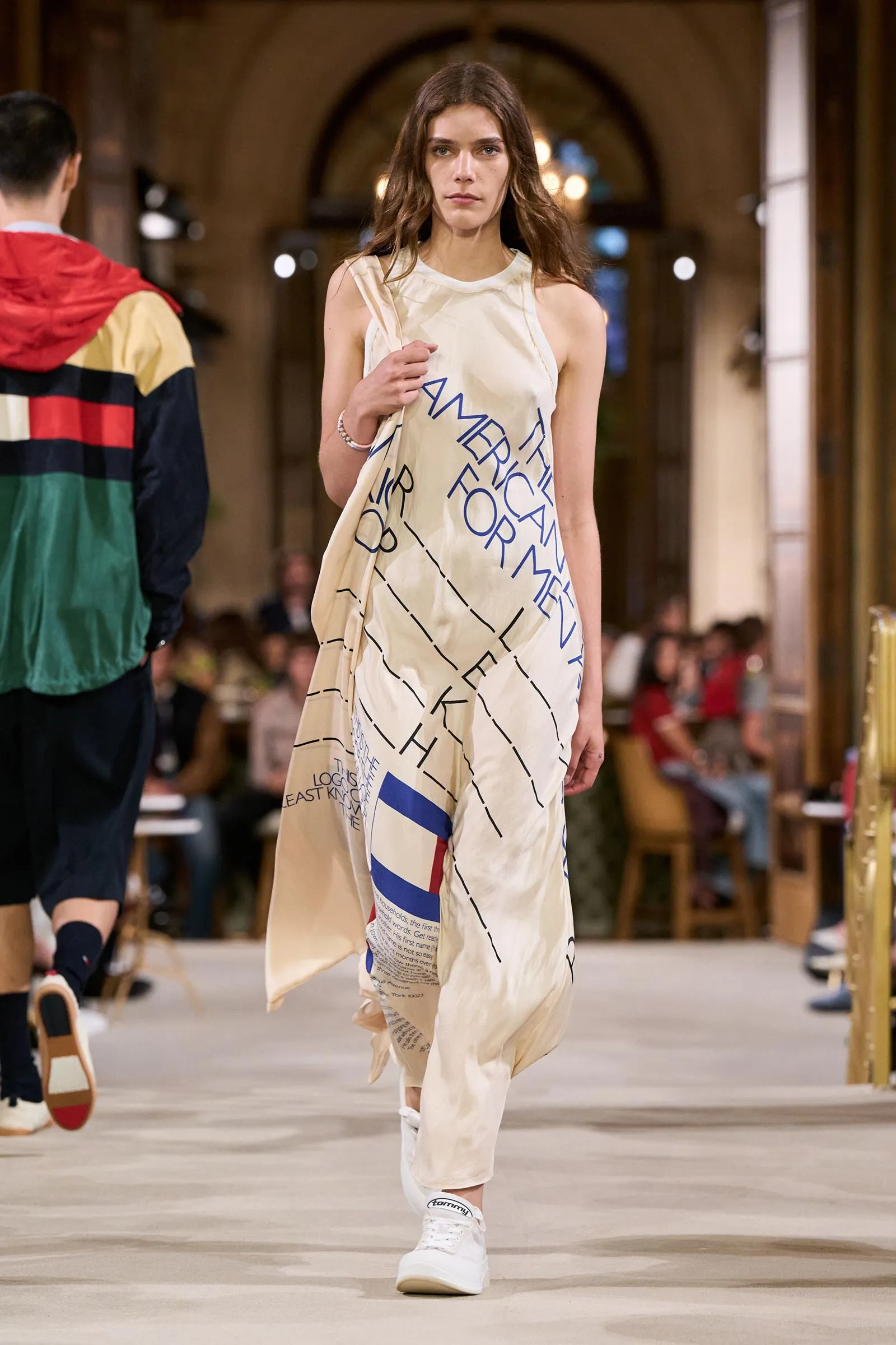 Model walking down a runway in a cream draped dress with blue typography, audience blurred in the background of a fashion show.