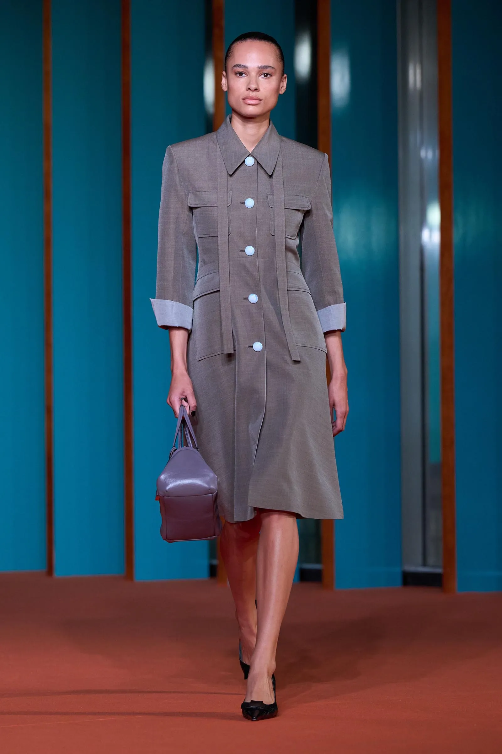 Model walking a runway in a gray trench dress with large white buttons, rolled cuffs, and a lavender bag, against teal panel backdrop.