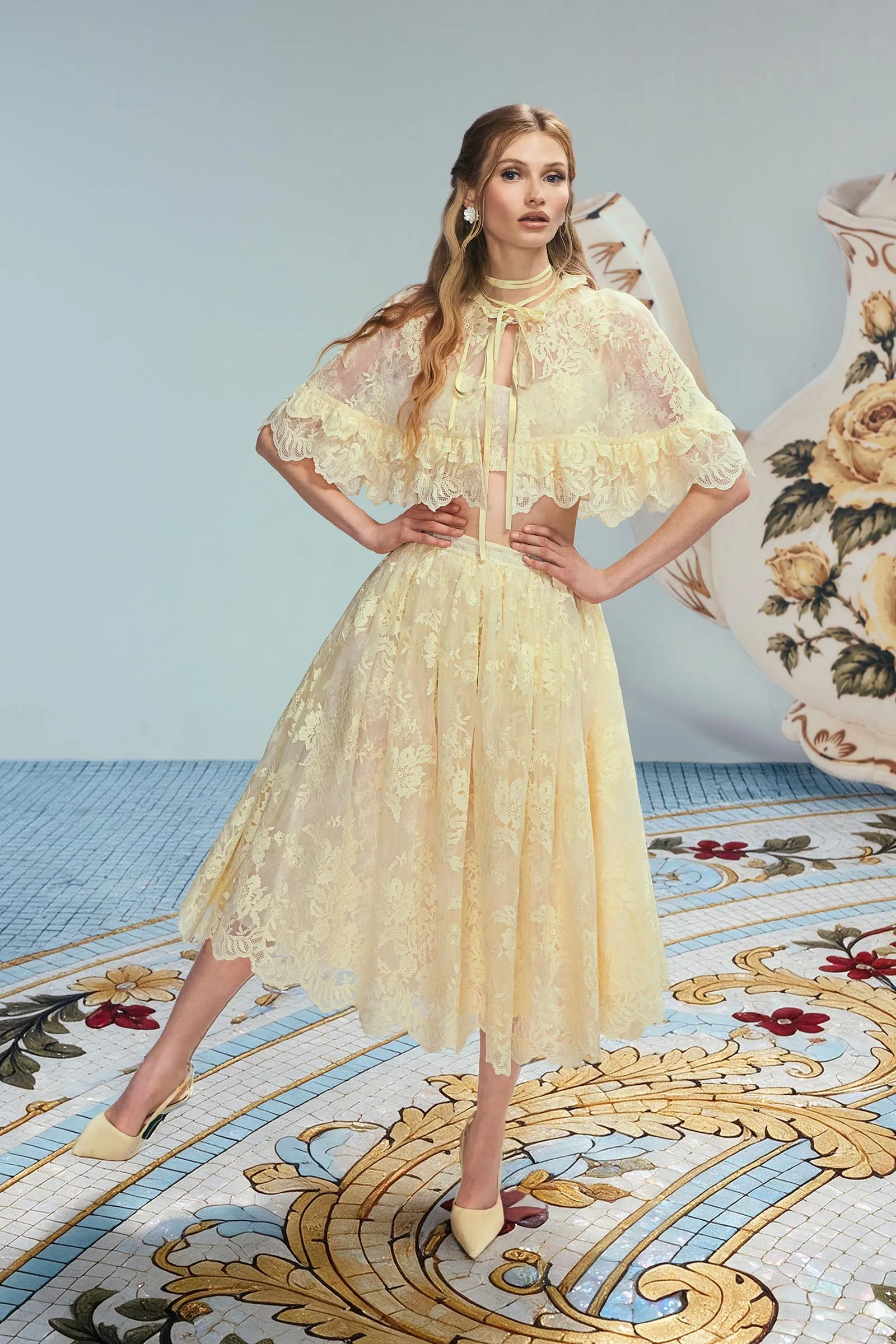 Model wearing a pale yellow lace dress with a sheer, ruffled cape, standing with hands on hips against a light blue wall and floral-patterned floor.