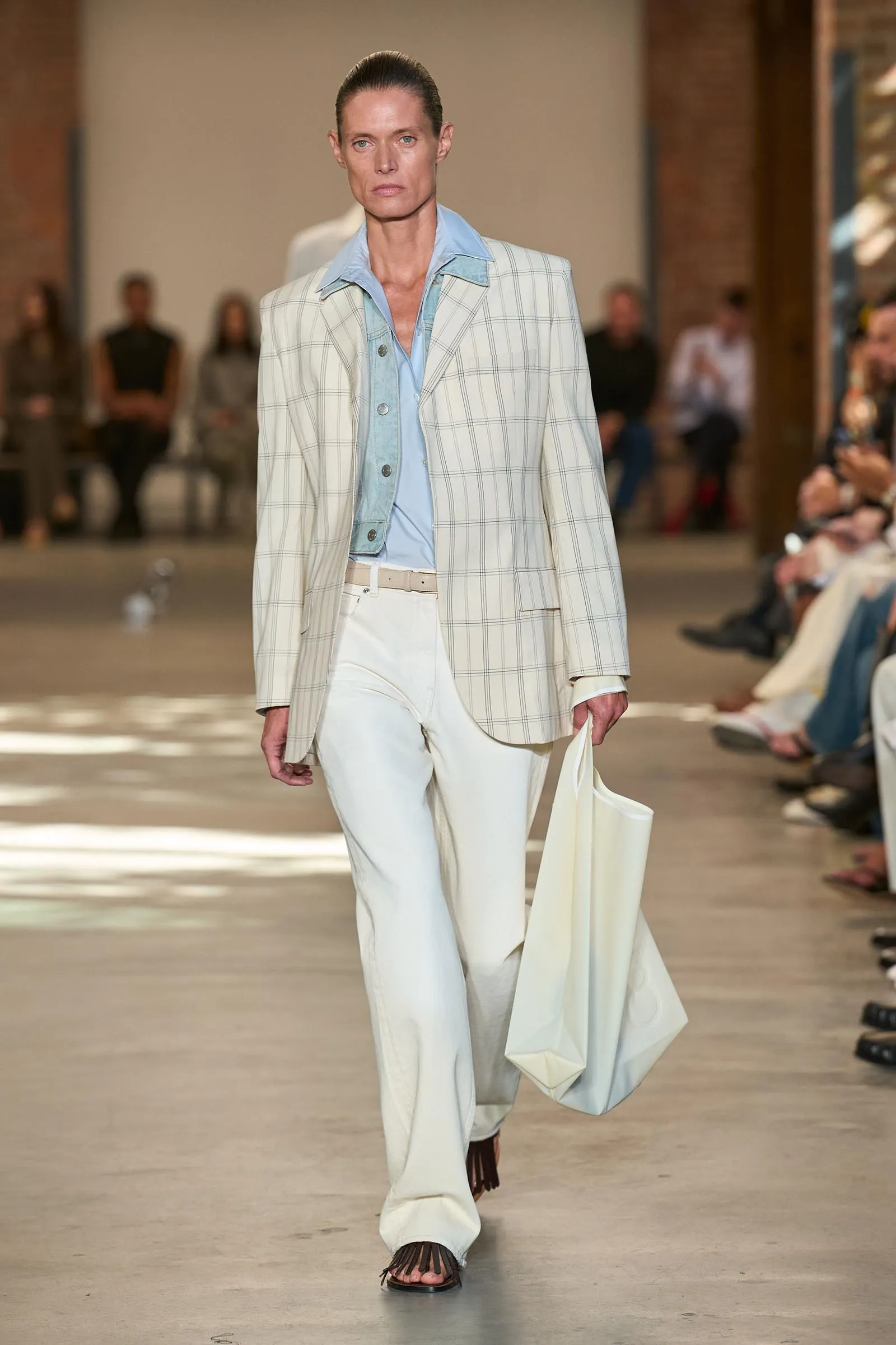 Model walks runway in a pale blue shirt under a cream plaid blazer with white wide-leg pants, carrying a white tote bag.