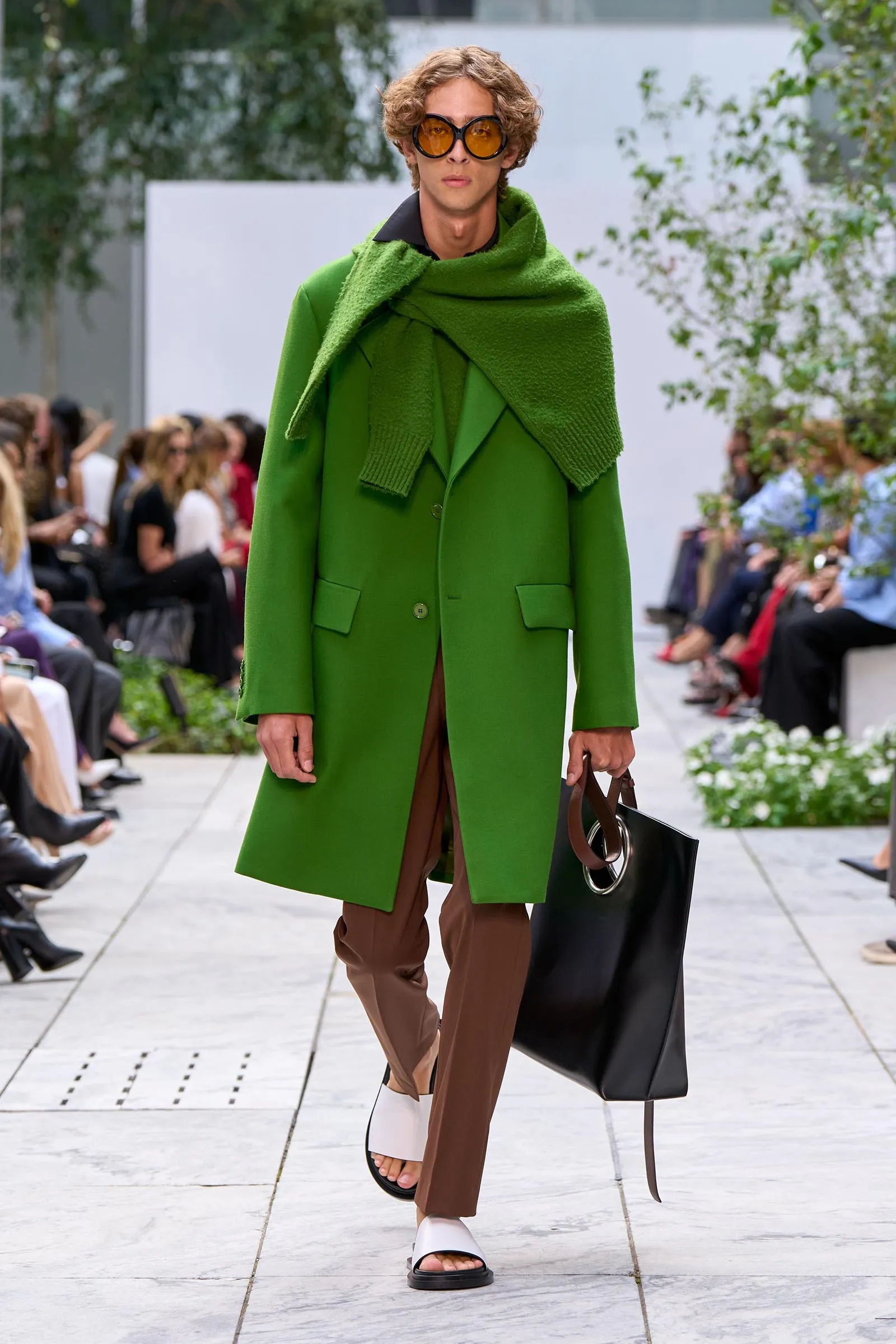 Model walking a runway in a vivid green coat with an oversized knit scarf, brown pants, white sandals, and a large black tote, audience in background.