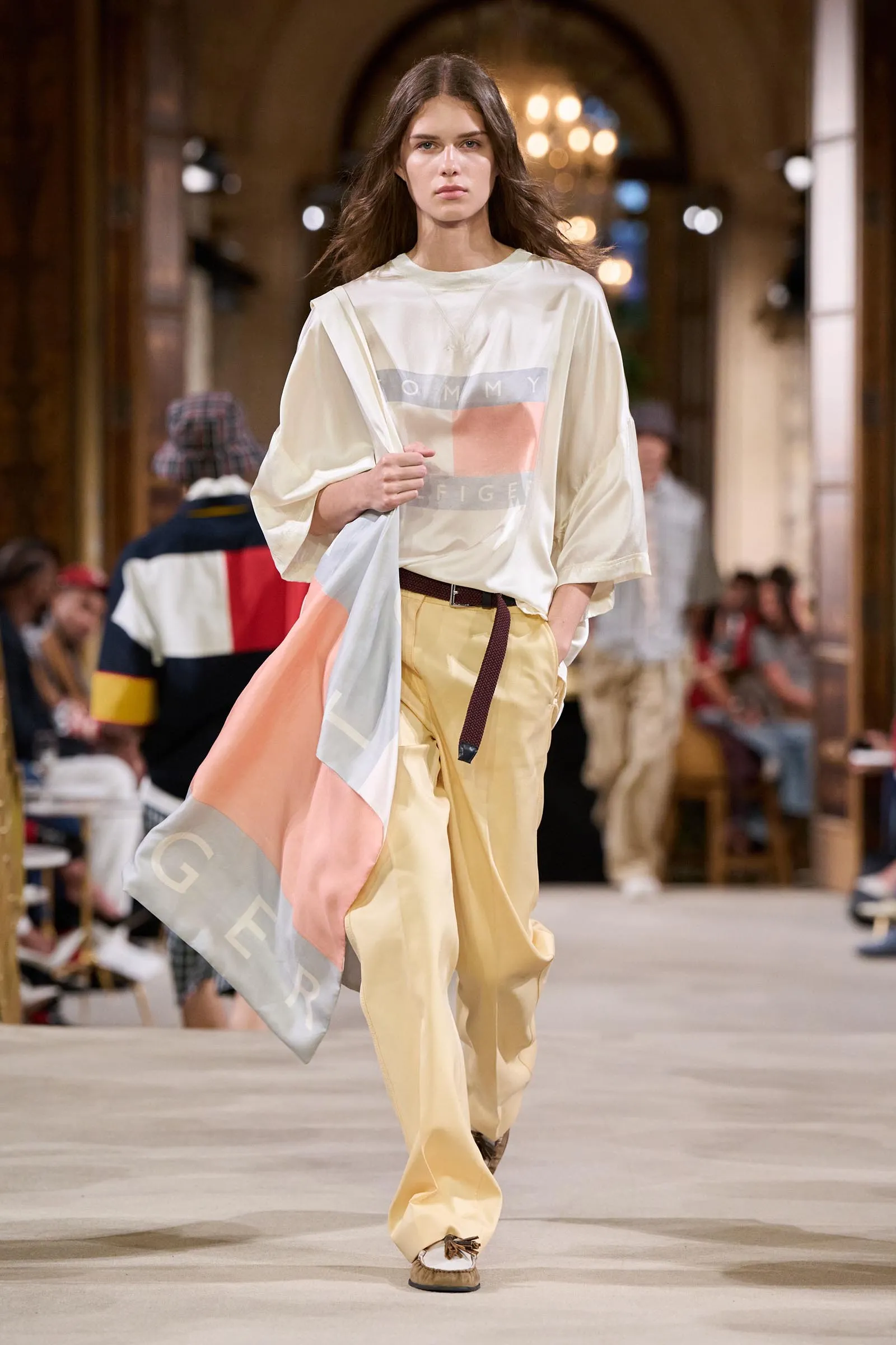 Model walking a runway in pastel-toned oversized top and tan pants, holding a draped scarf while audience watches in a grand venue.