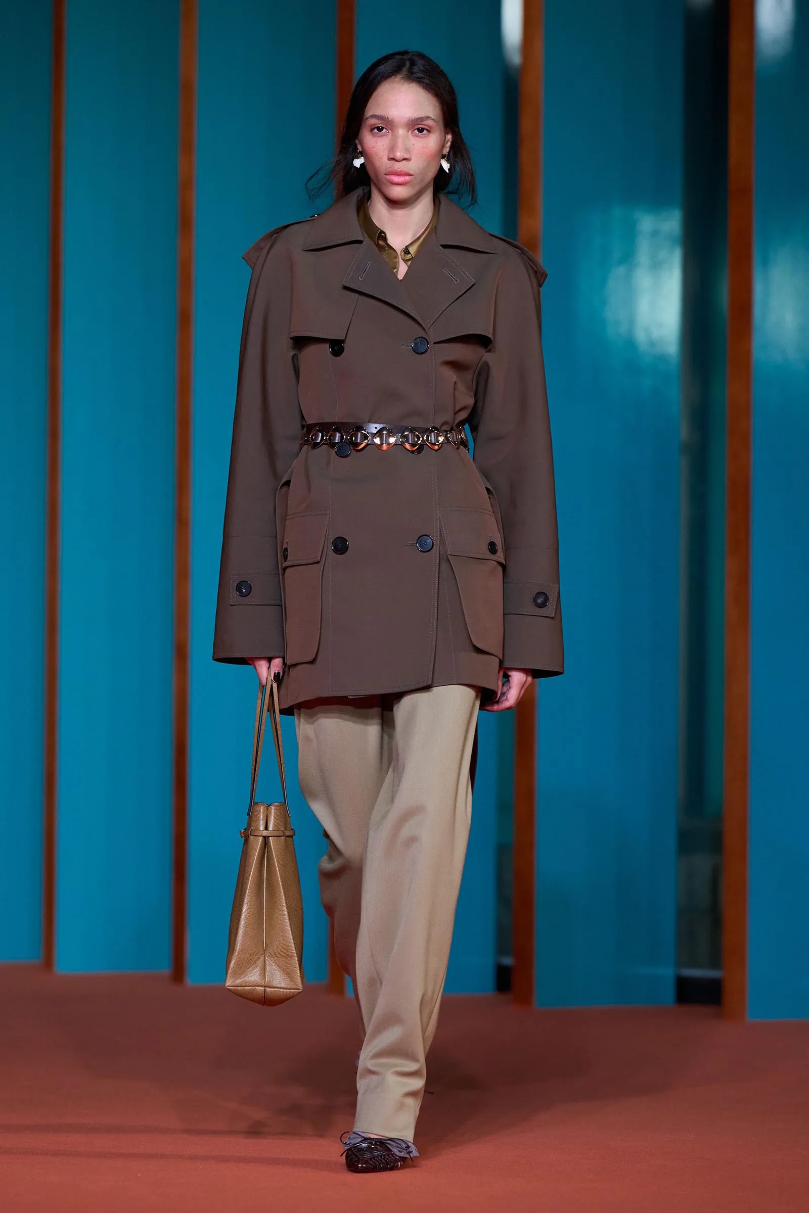 Female model walks the runway in a brown belted trench coat with beige trousers, carrying a tan handbag.