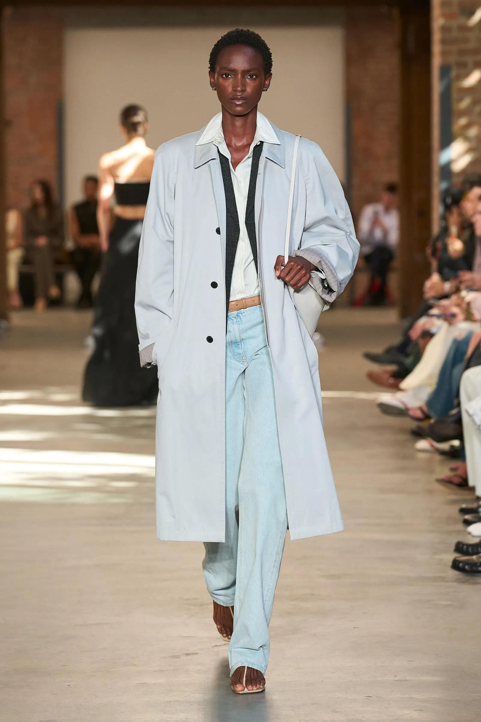 Female model walks a fashion runway in a pale blue trench coat over a white shirt and light denim jeans, sandals, with spectators in the background.