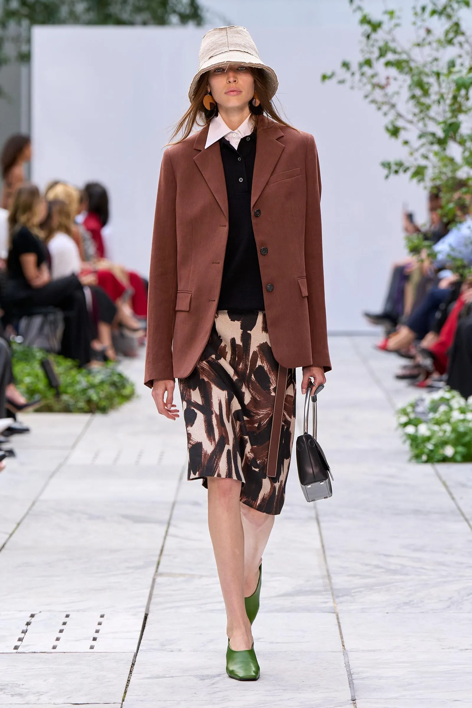 Model walking the runway in a brown blazer over a black polo with a patterned midi skirt and green shoes, holding a small handbag.