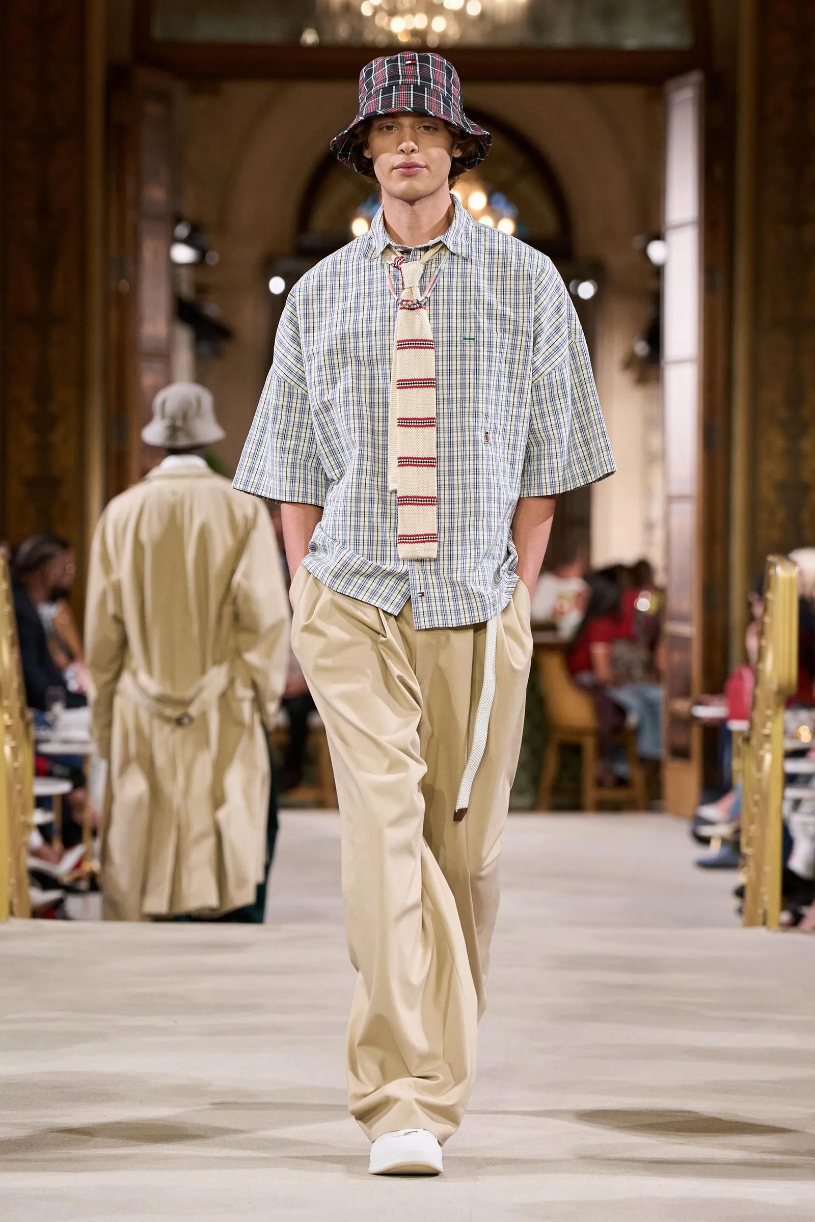 Male model walking a runway in a plaid bucket hat, loose checkered shirt, beige tie and khaki pants in a fashion show setting.