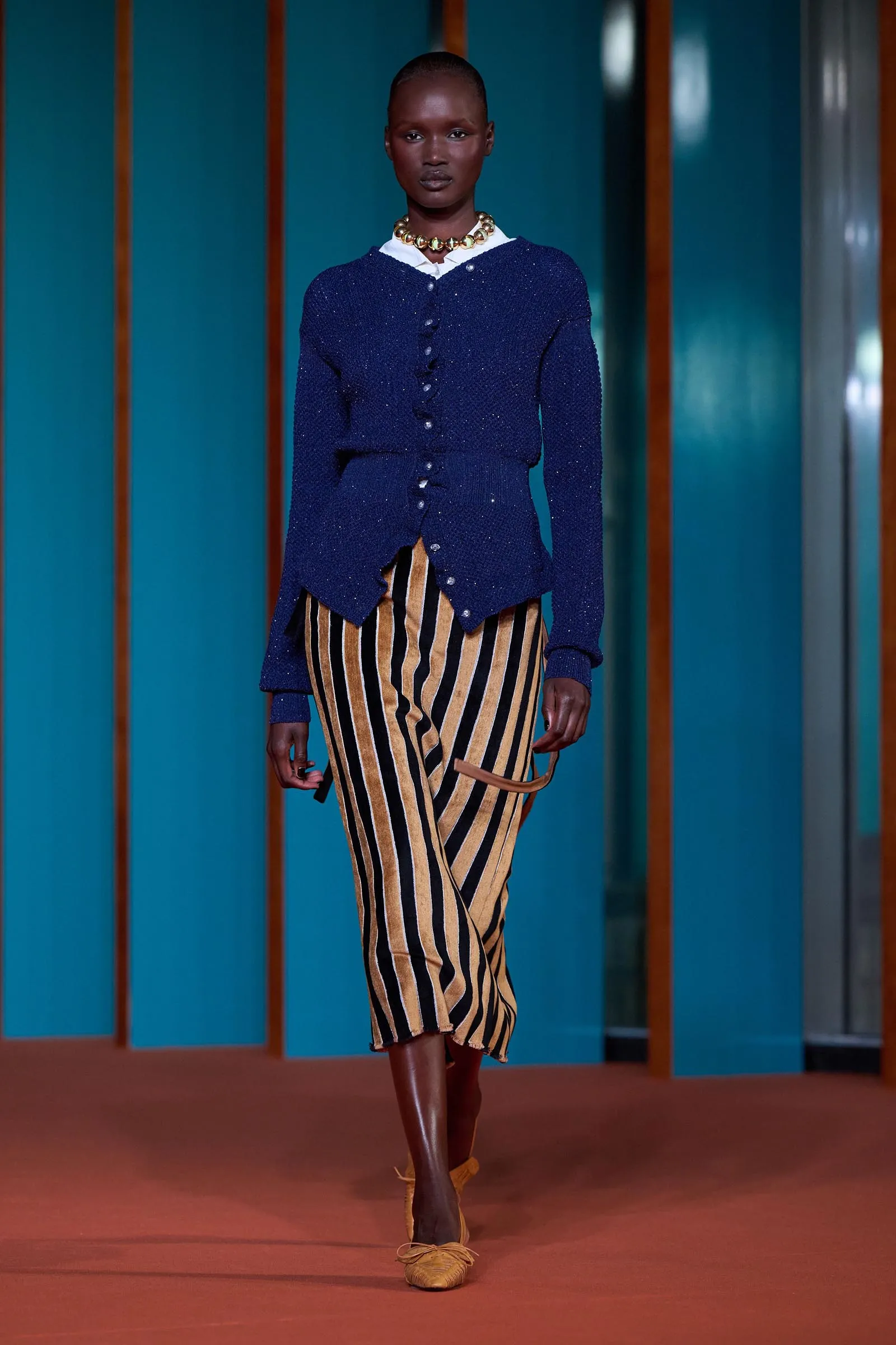 Model walking on a runway wearing a navy blue knit cardigan with a white collar and a gold striped skirt, accessorized with a chunky bead necklace.