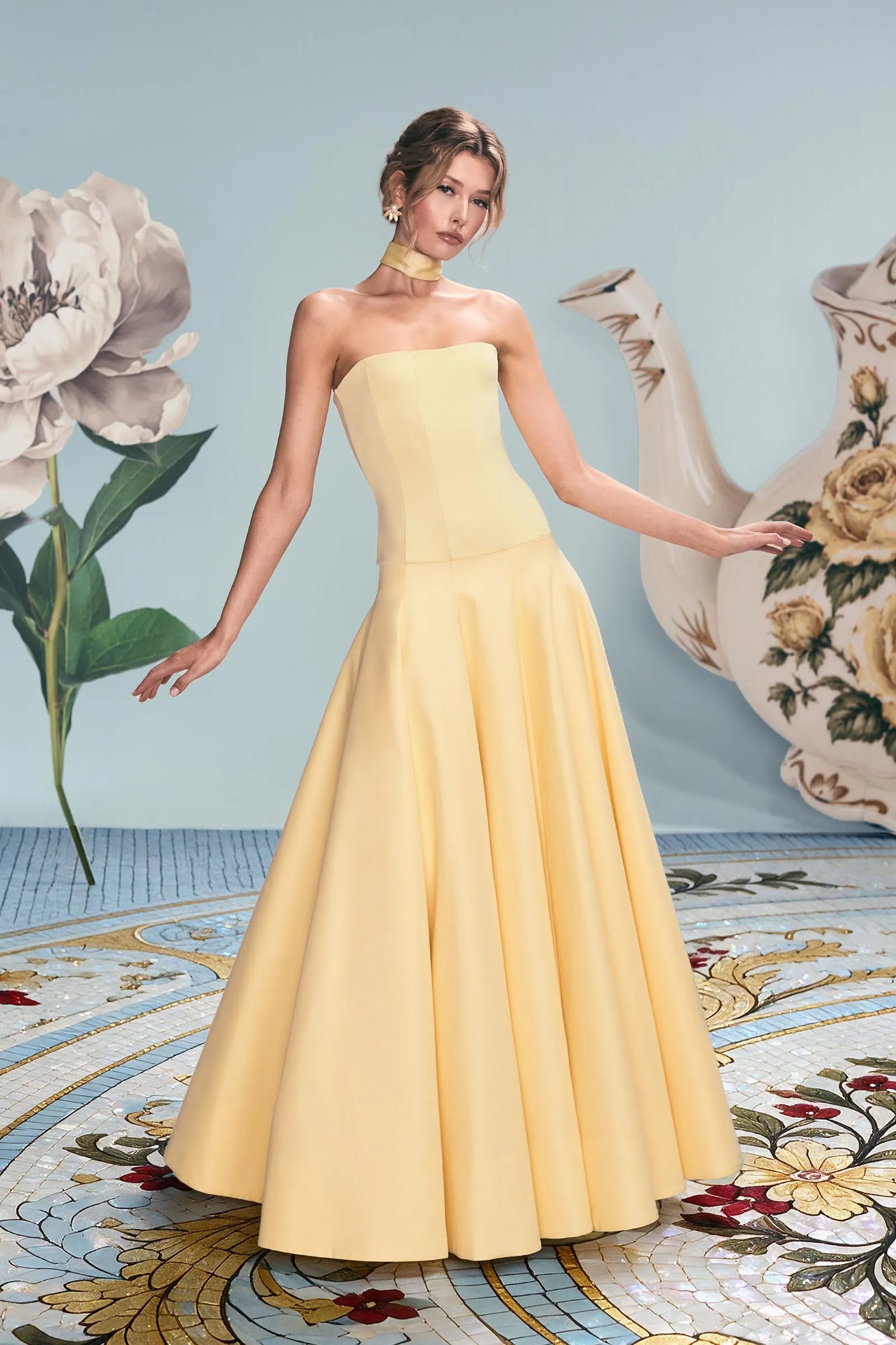 Model in a pale yellow strapless gown and choker necklace poses with arms outstretched in a pastel studio with oversized white flowers and a decorative teapot sculpture in the background.