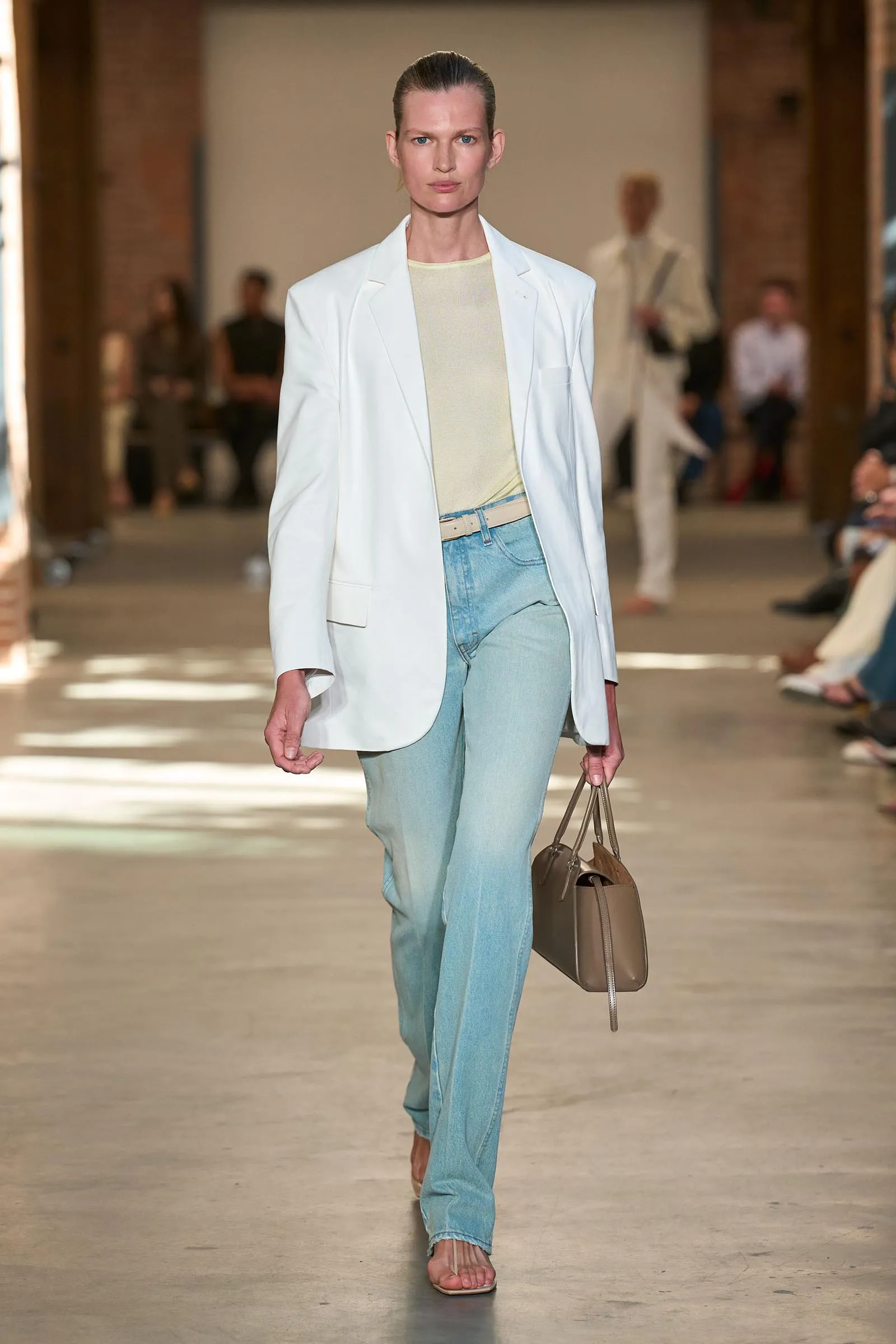 Model walks runway in a white blazer over a pale sweater and light-wash jeans, carrying a taupe handbag.