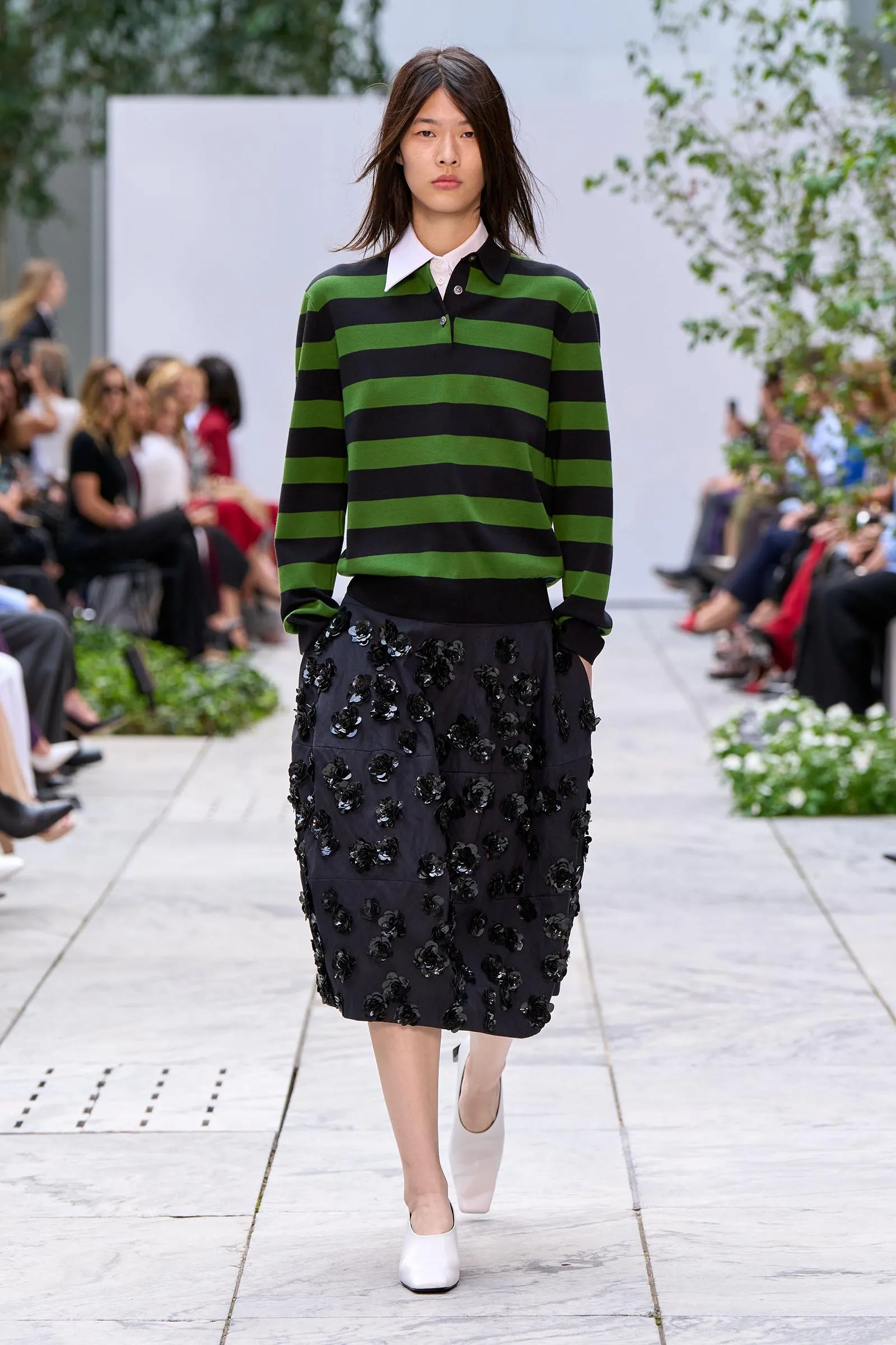 Model walking the runway in a green and black horizontal striped sweater with a dark skirt covered in black floral appliqués.
