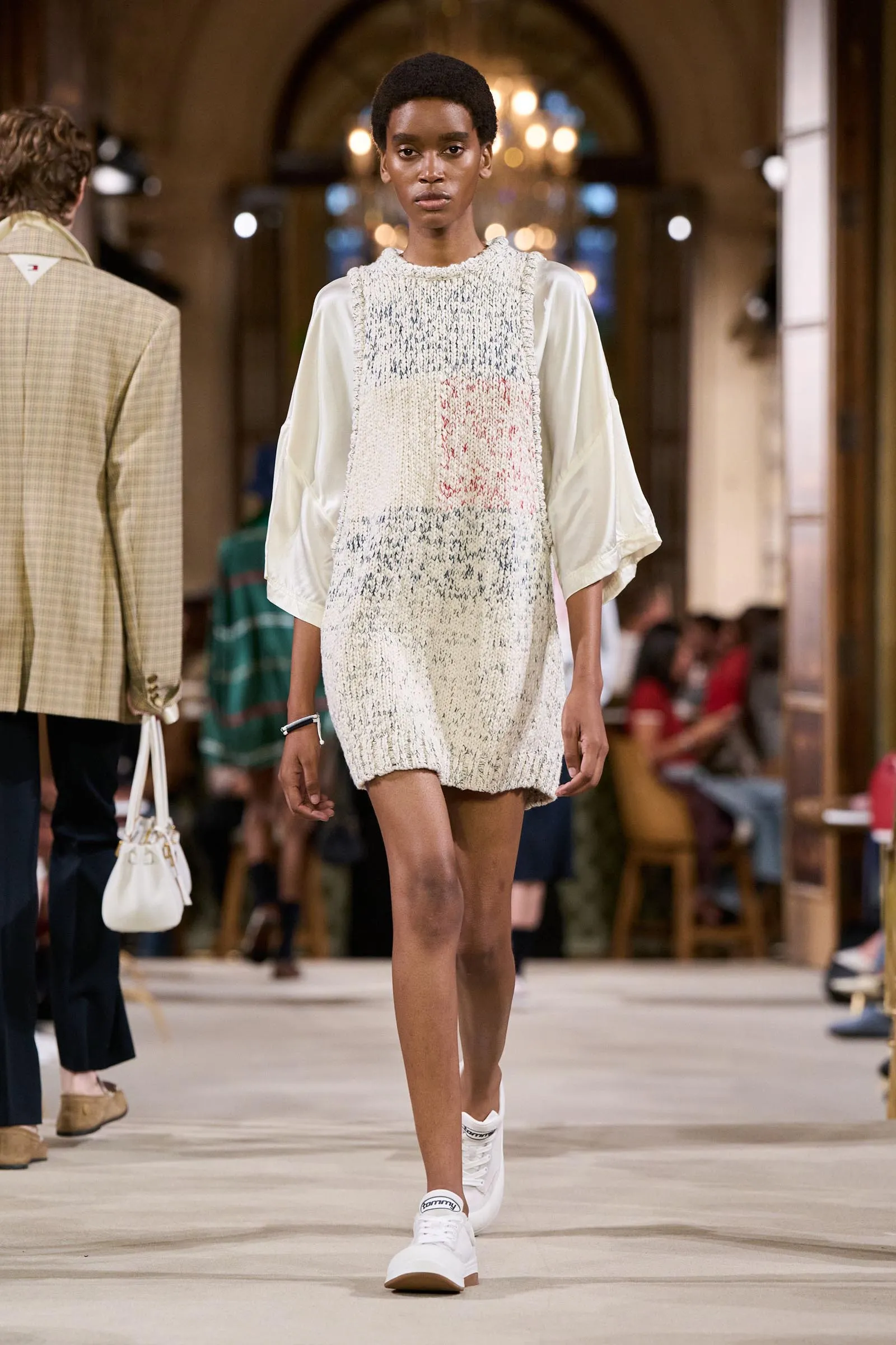Model walks the runway wearing a cream textured knit sleeveless dress over a pale blouse, with white sneakers.