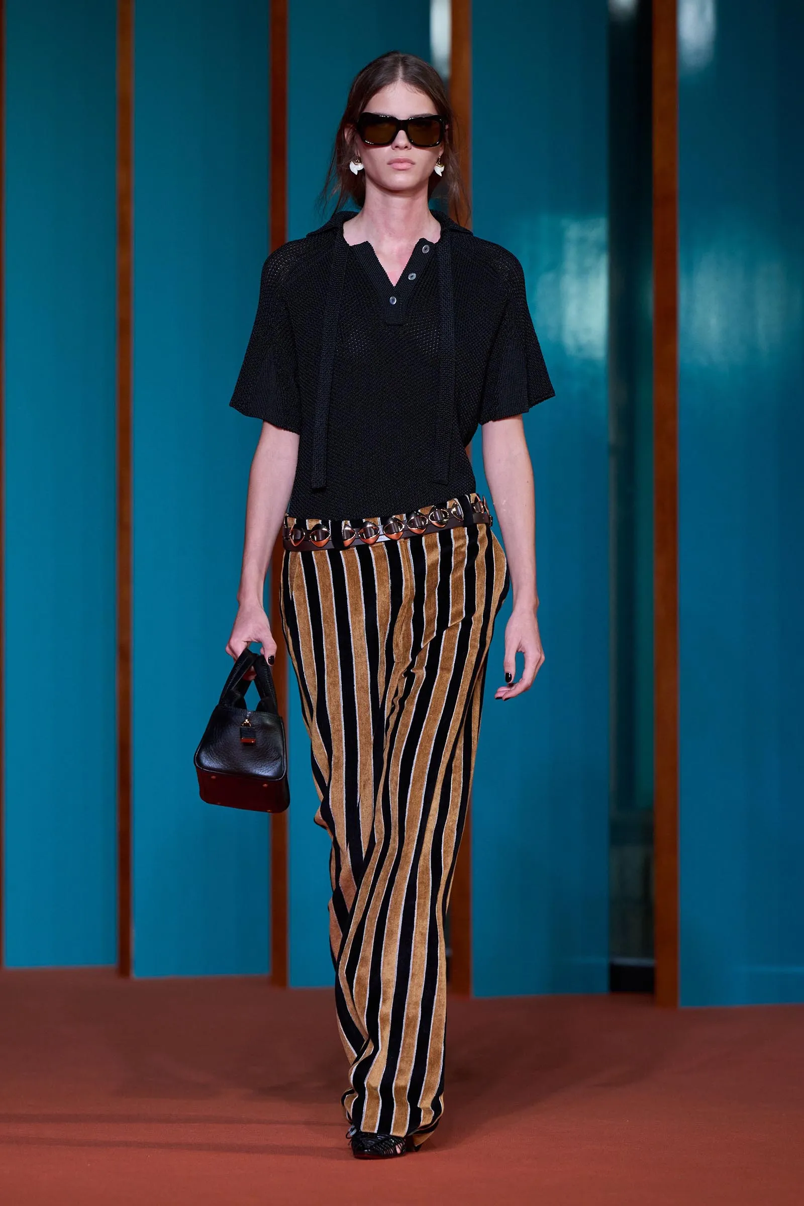 Model walking on a runway in a black knit top and vertically striped brown-and-black pants, carrying a small black handbag and wearing oversized sunglasses.