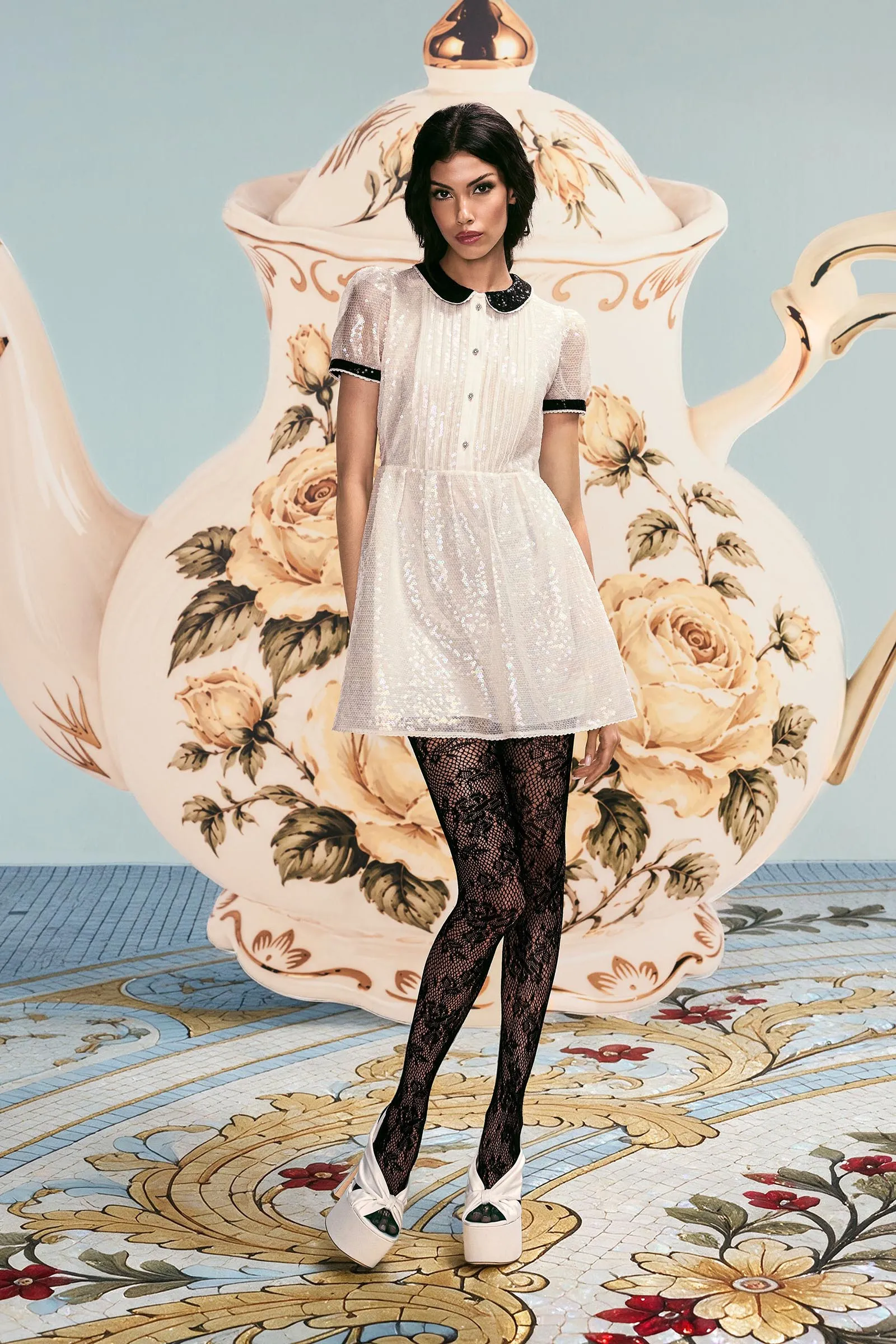 Model in a silver sequin dress with black collar, black lace tights, and white platform shoes, posing before a large floral teapot backdrop.