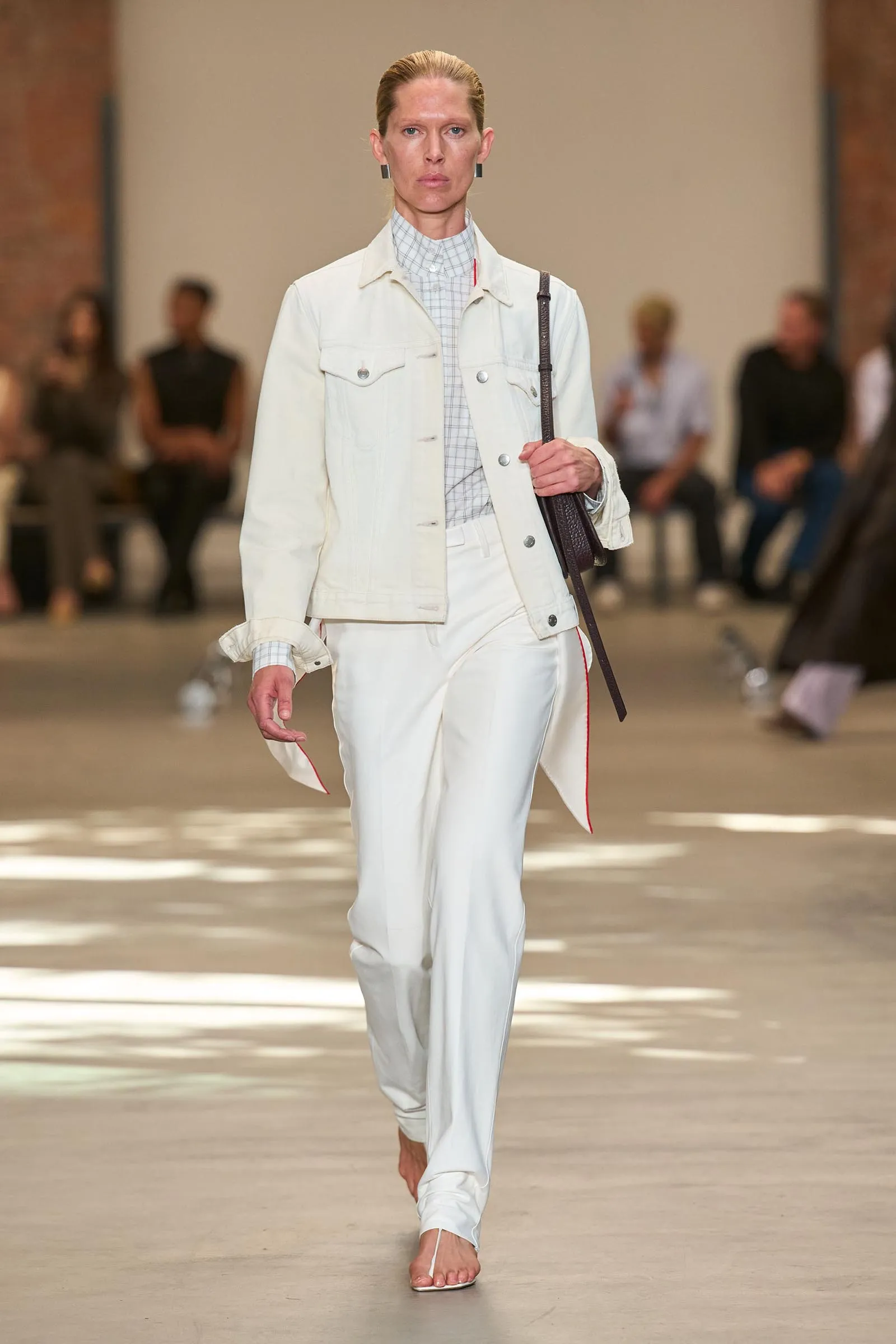 Model walking a fashion runway in a pale white denim jacket over a light checkered shirt and matching white pants, carrying a dark brown shoulder bag.