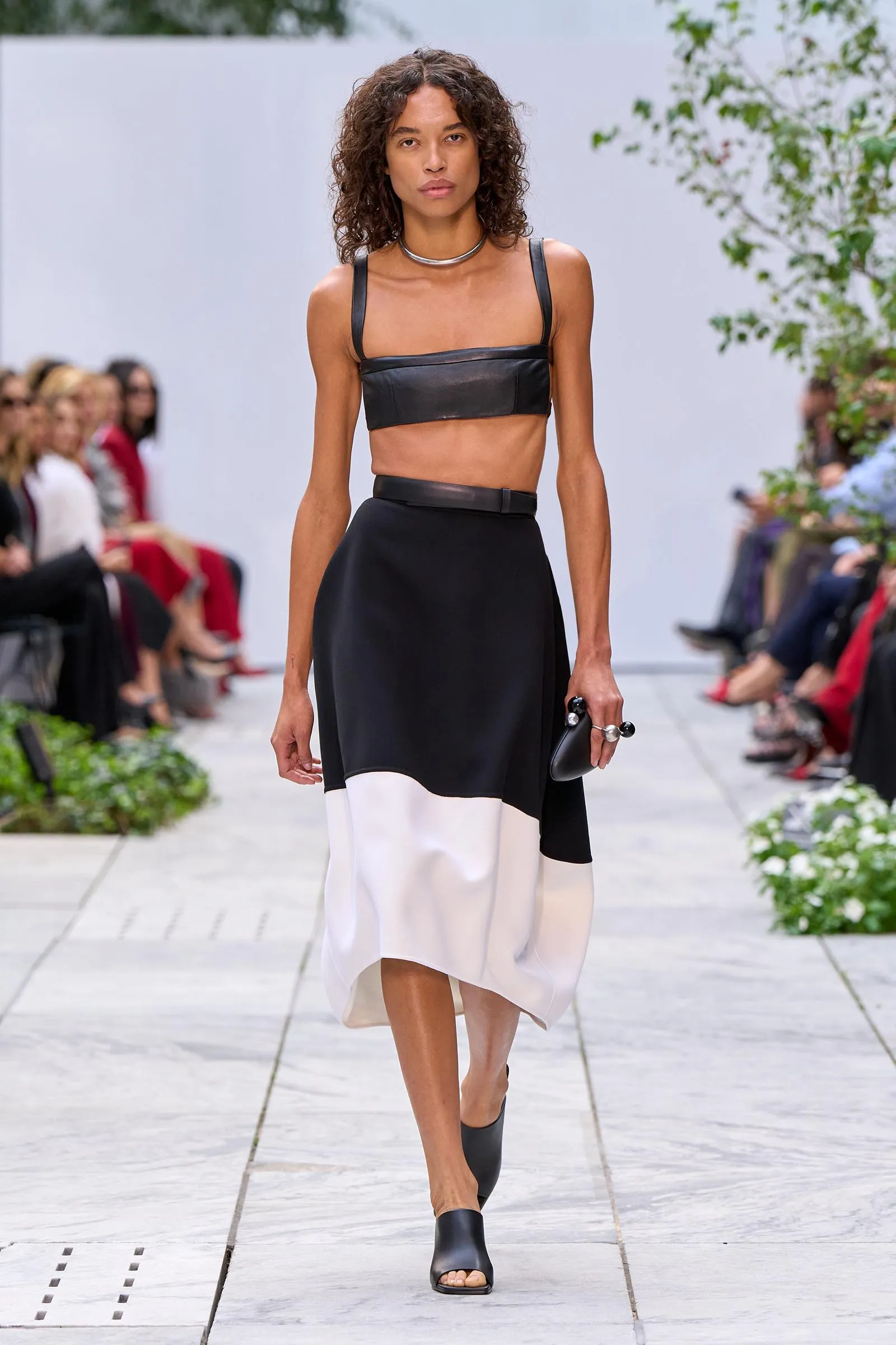 Model walks a fashion runway wearing a black leather bandeau top with matching belt and a color-block black-and-white skirt, accessorized with black open-toe sandals.