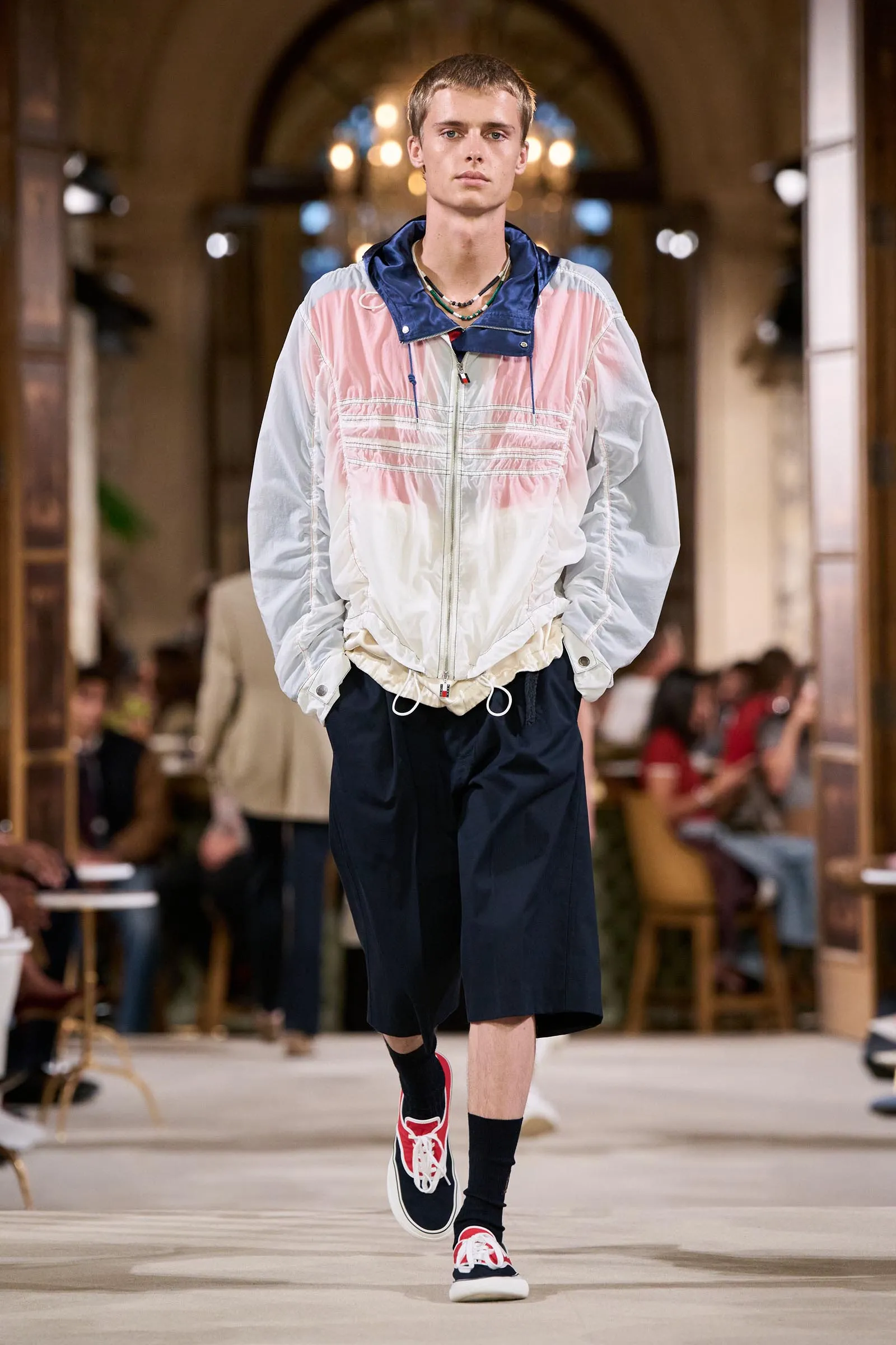 Male model walks a fashion-show runway in a pastel pink-and-white windbreaker, navy shorts, and red sneakers, with an audience in the background.