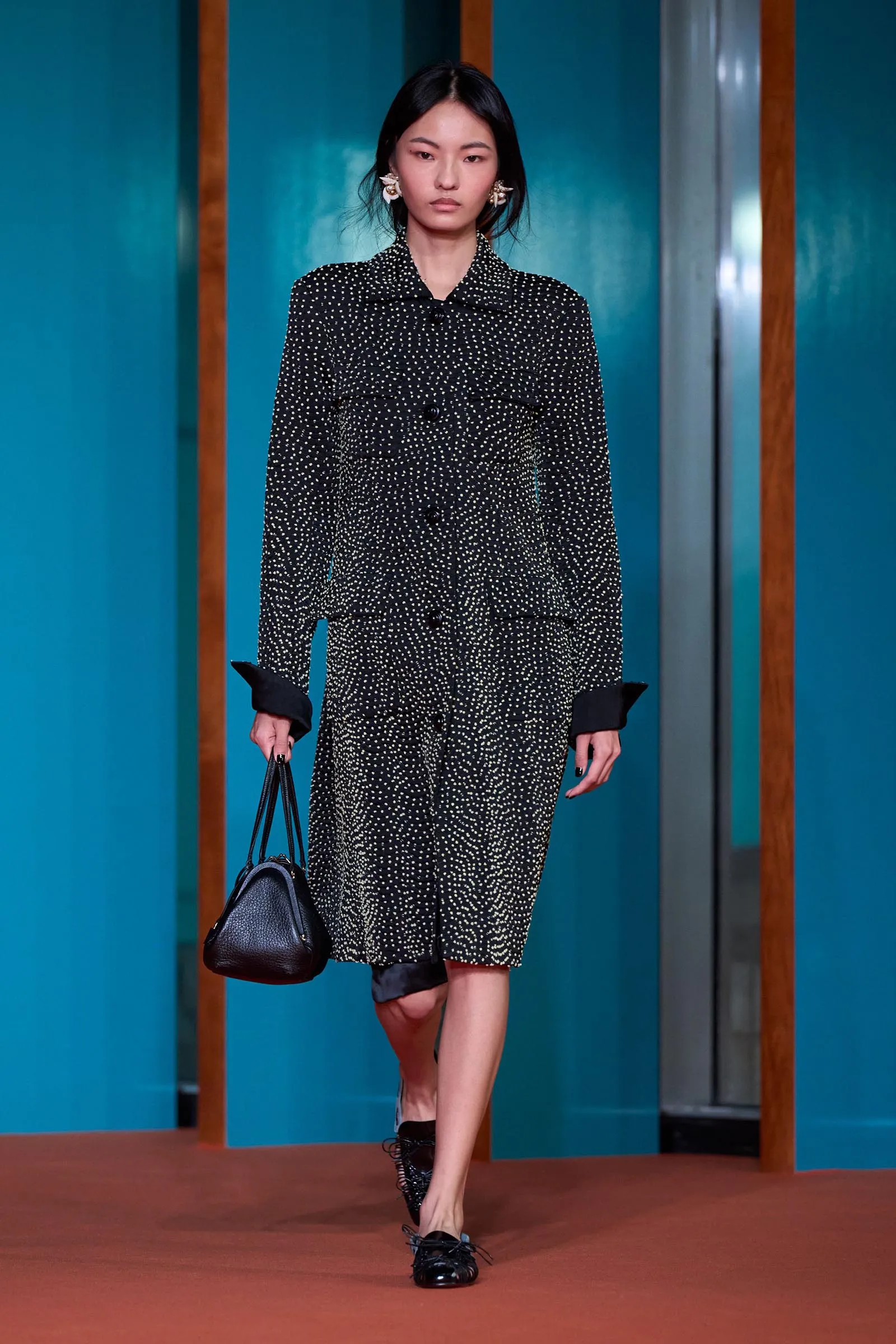 Model walking a runway in a black polka-dot suit with rolled cuffs, carrying a black handbag against teal walls with wooden vertical accents.