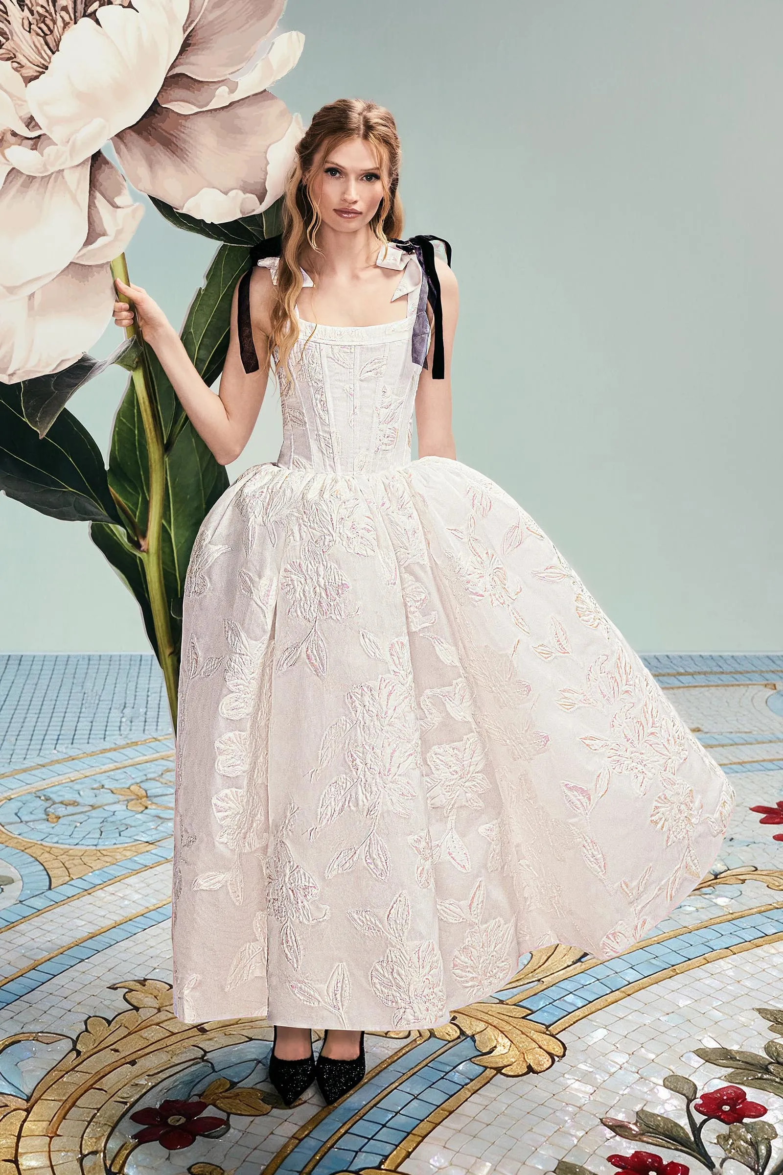 Woman in a pale pink, embroidered gown posing with an oversized flower prop and green leaves, standing on a decorative mosaic floor with pale blue background.