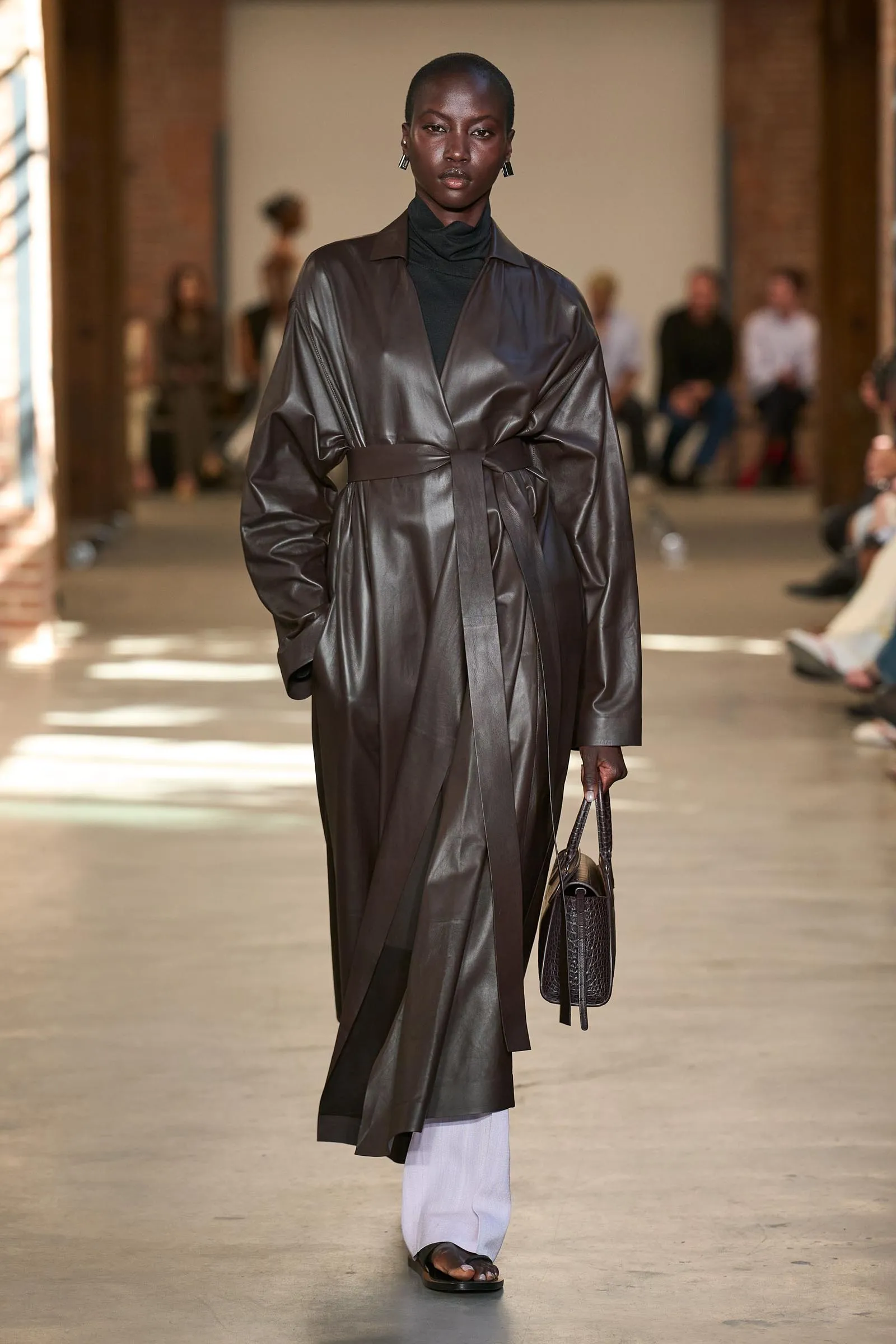 Runway model in a shiny charcoal-gray trench coat belted at the waist, black turtleneck, pale pants, and a dark textured handbag as she walks toward the camera.