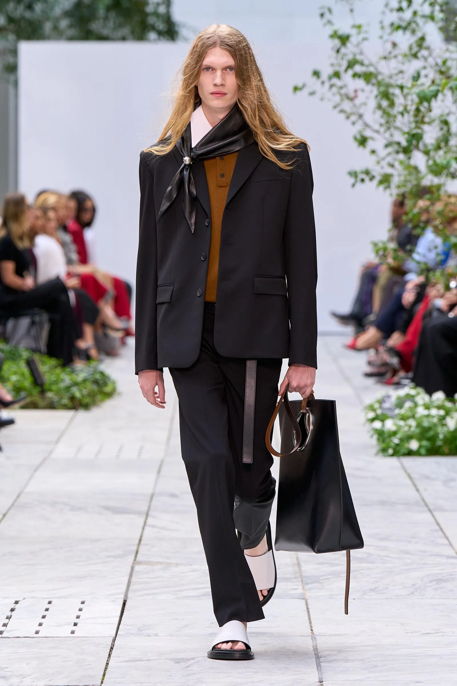 Model walking a runway in a black suit with a brown top, accessorized by a black scarf and a black tote bag.