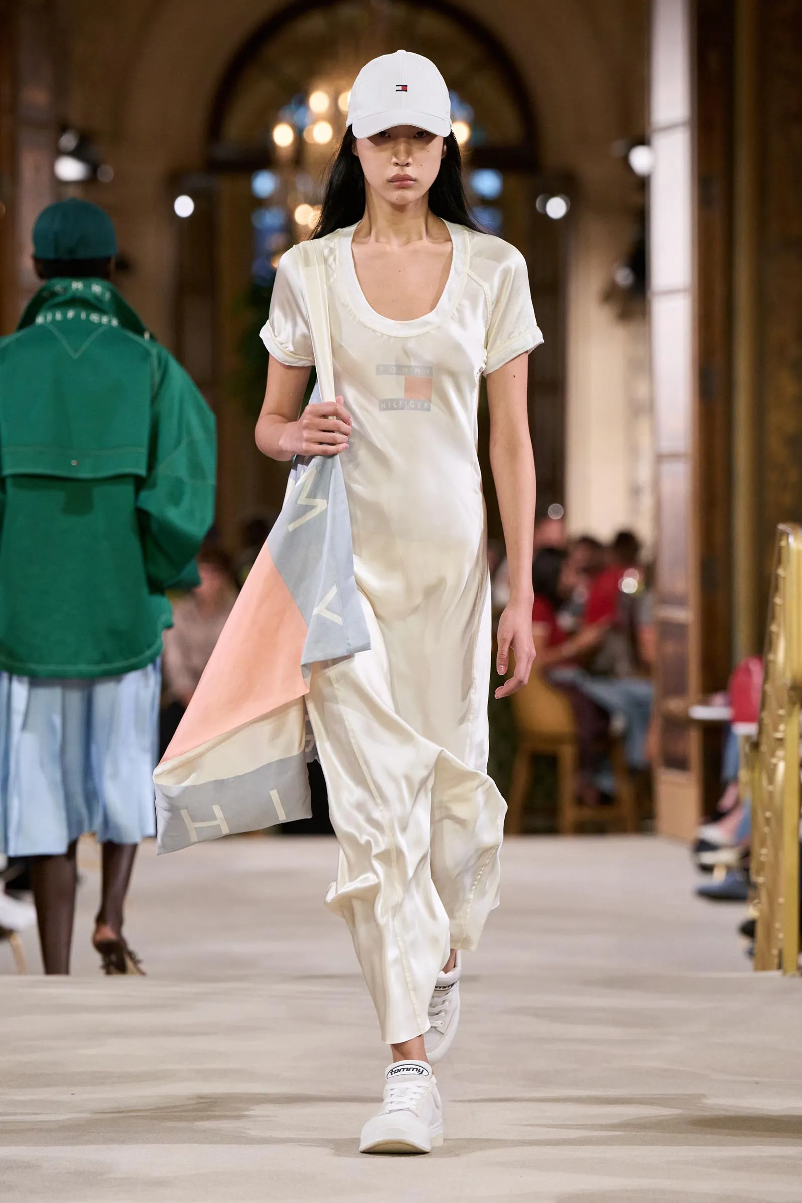 Model walking a runway in a light cream dress and white cap, carrying a pastel scarf.