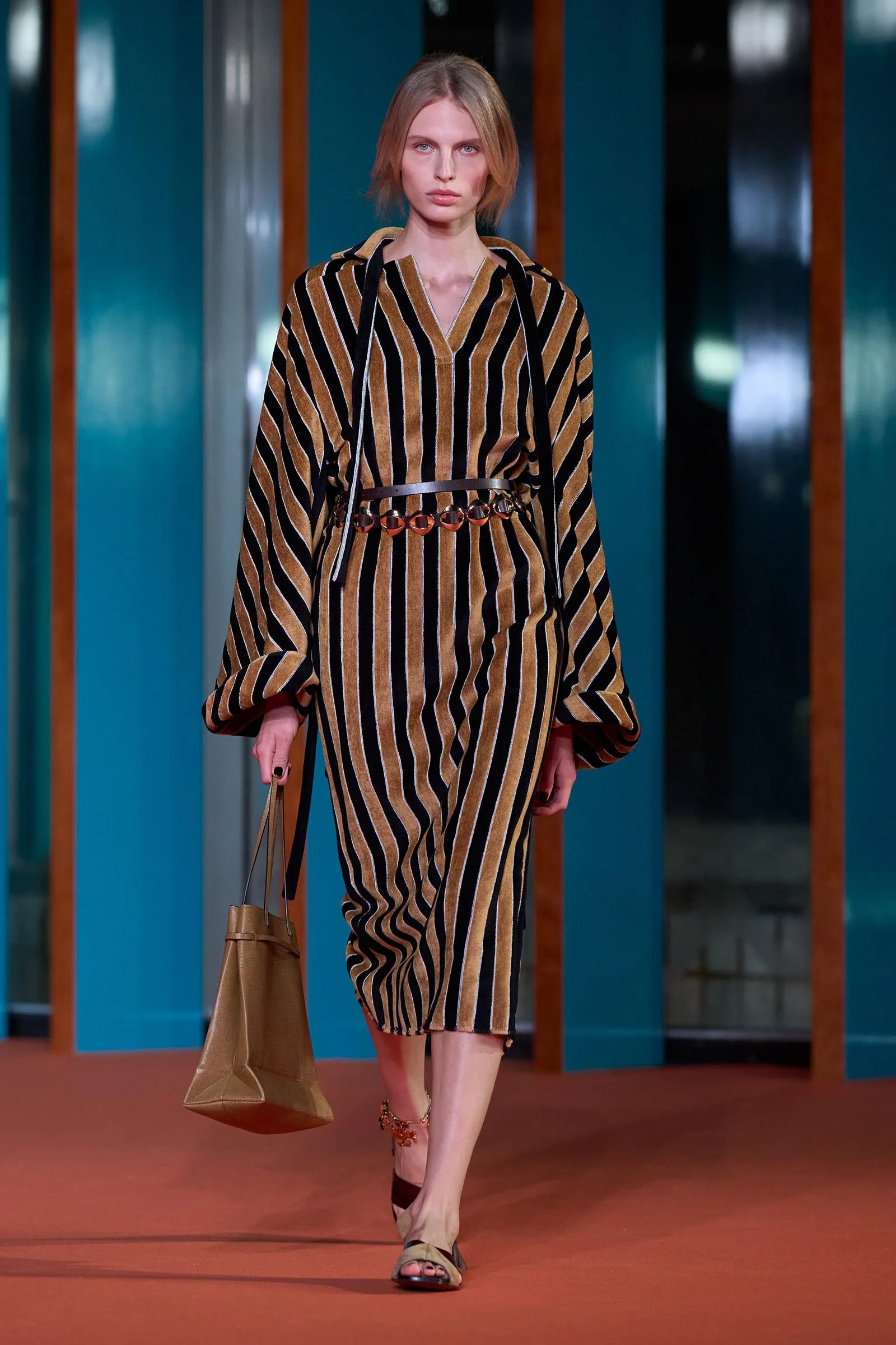 Model walking the runway in a brown-and-black striped velvet dress with a brown belt and beige handbag
