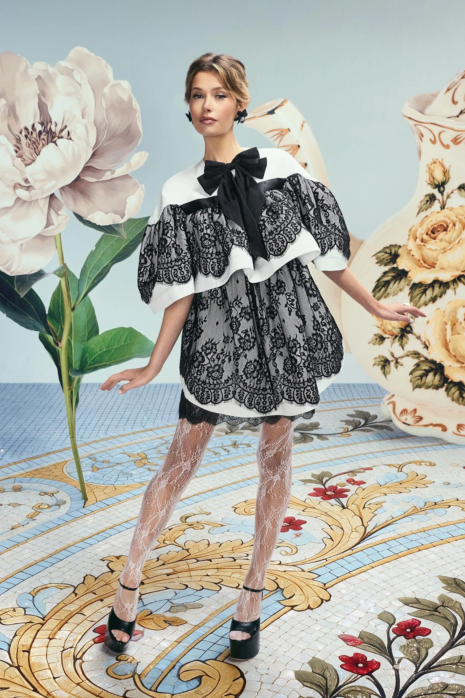 Model wearing a black lace dress with a white yoke and large bow, posing in a floral-studio set with oversized flowers in the background and patterned tiled floor.