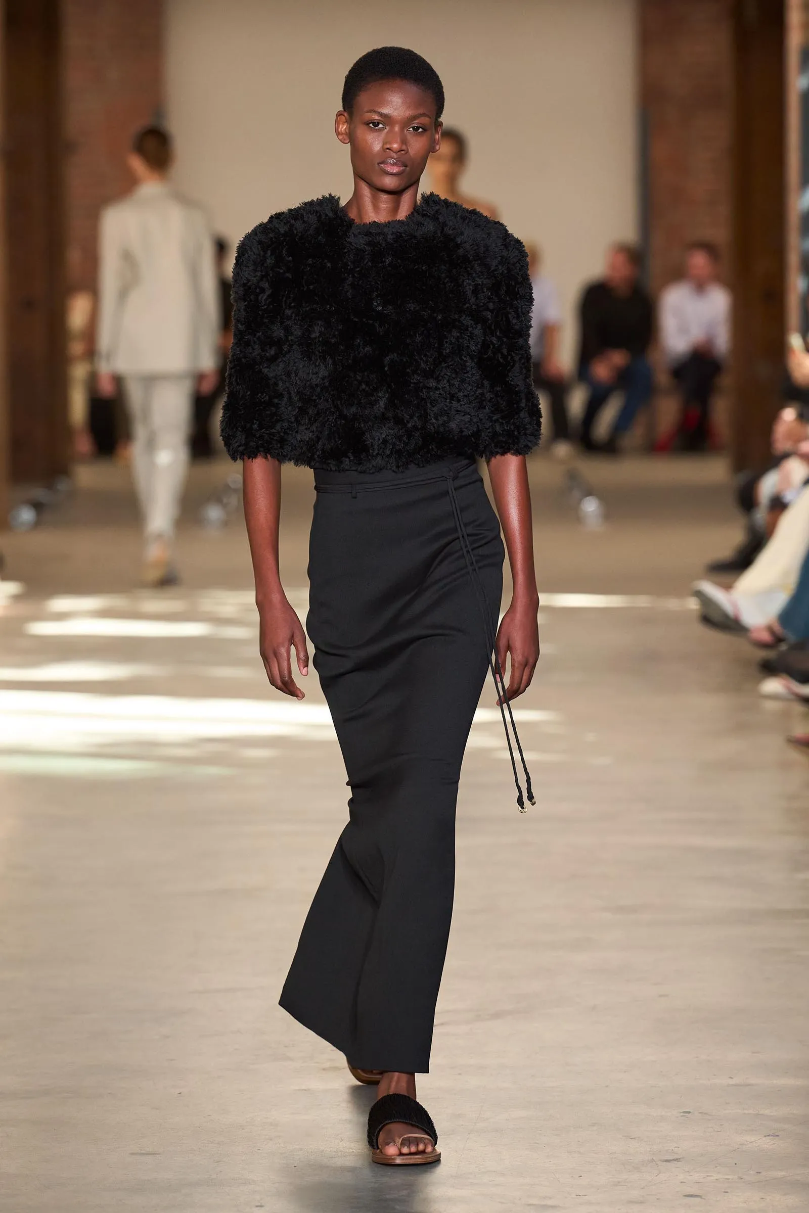 Model walks a fashion runway wearing a fluffy black textured top and black pants, in a studio setting with blurred audience in the background.
