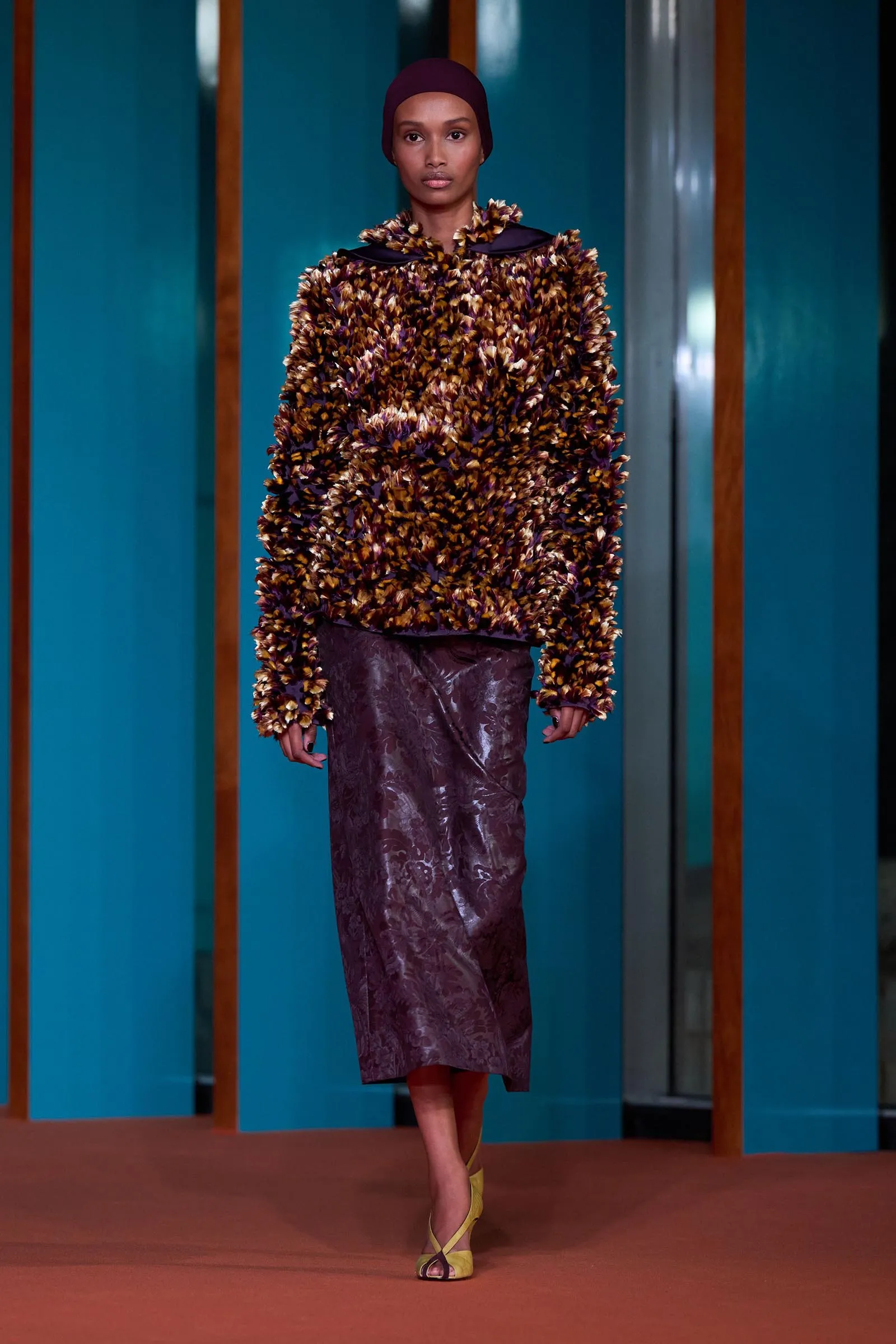 Model walking a fashion runway in a brown feathered top, purple skirt, and burgundy headwrap against teal panels.