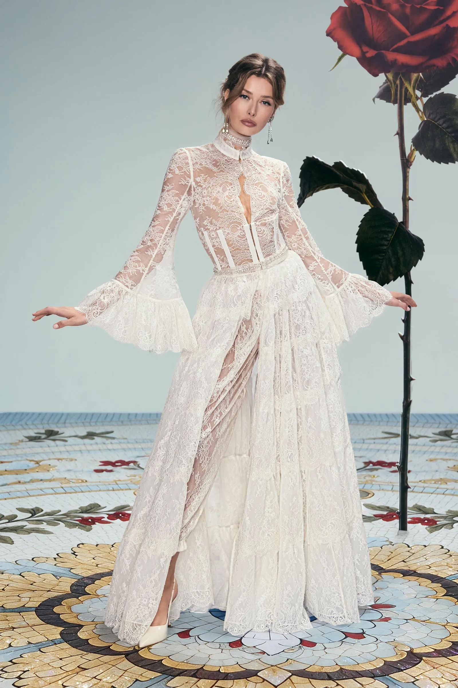 Model in an ivory lace wedding dress posing with outstretched arms beside a large red rose on a mosaic floor.