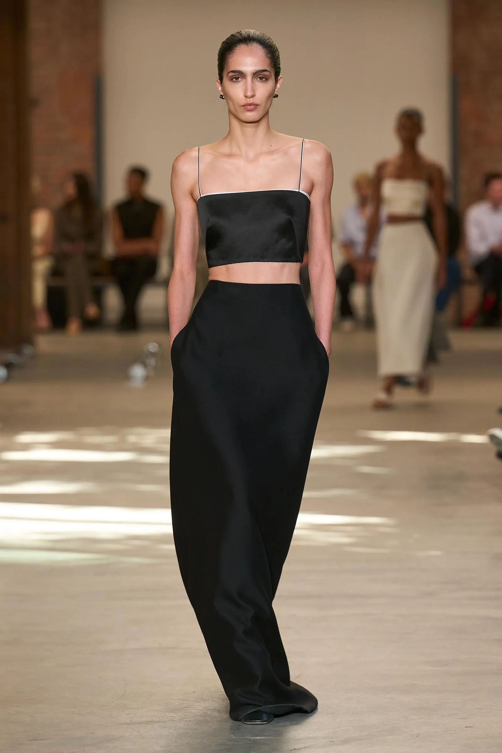 Model walks the runway in a black spaghetti-strap crop top and matching high-waisted skirt/pants combo. Negative space shows a blurred audience behind.