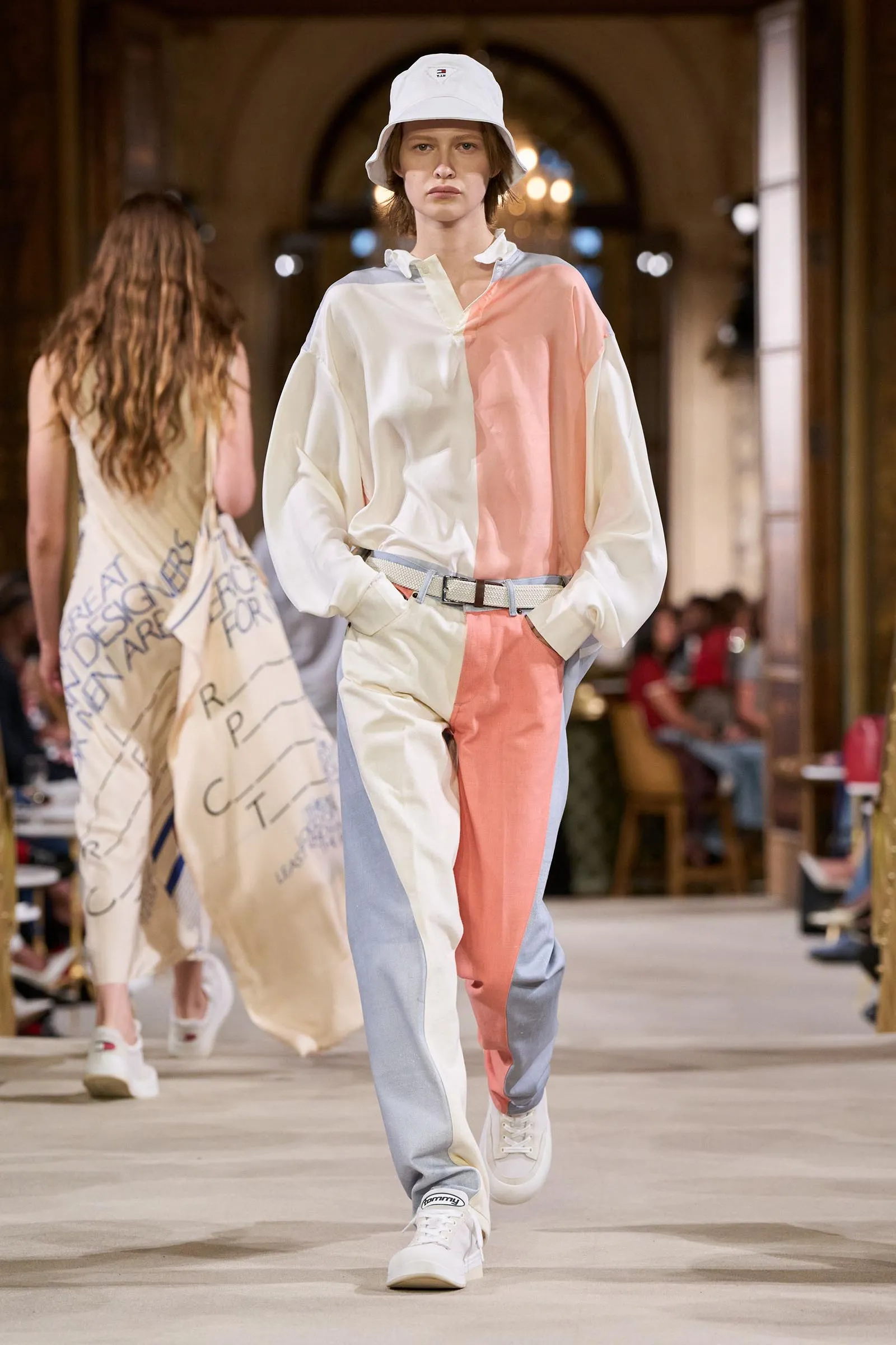 Model on a fashion runway wearing a pastel color-block jumpsuit and white bucket hat, walking toward the camera.