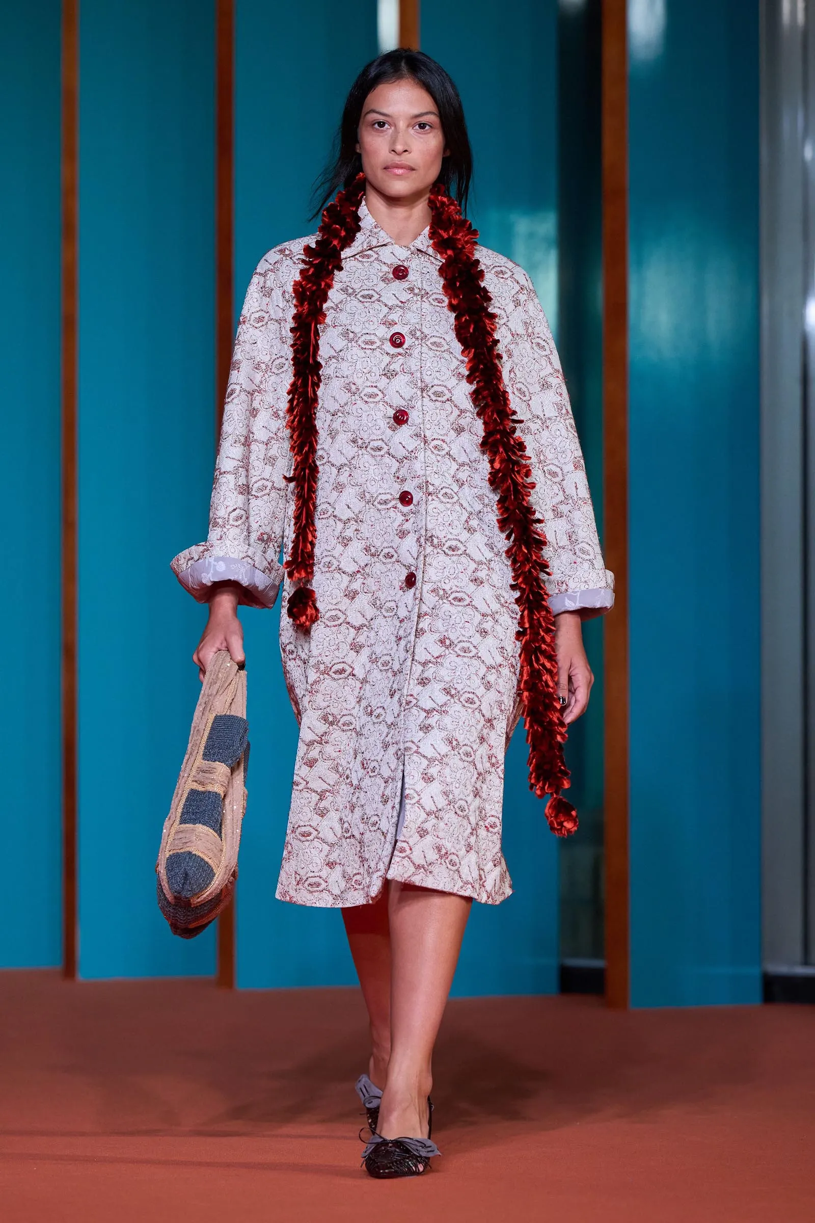 Woman walking a fashion runway in a white lace dress with red buttons and a red feathered garland, holding a woven bag against teal panels.