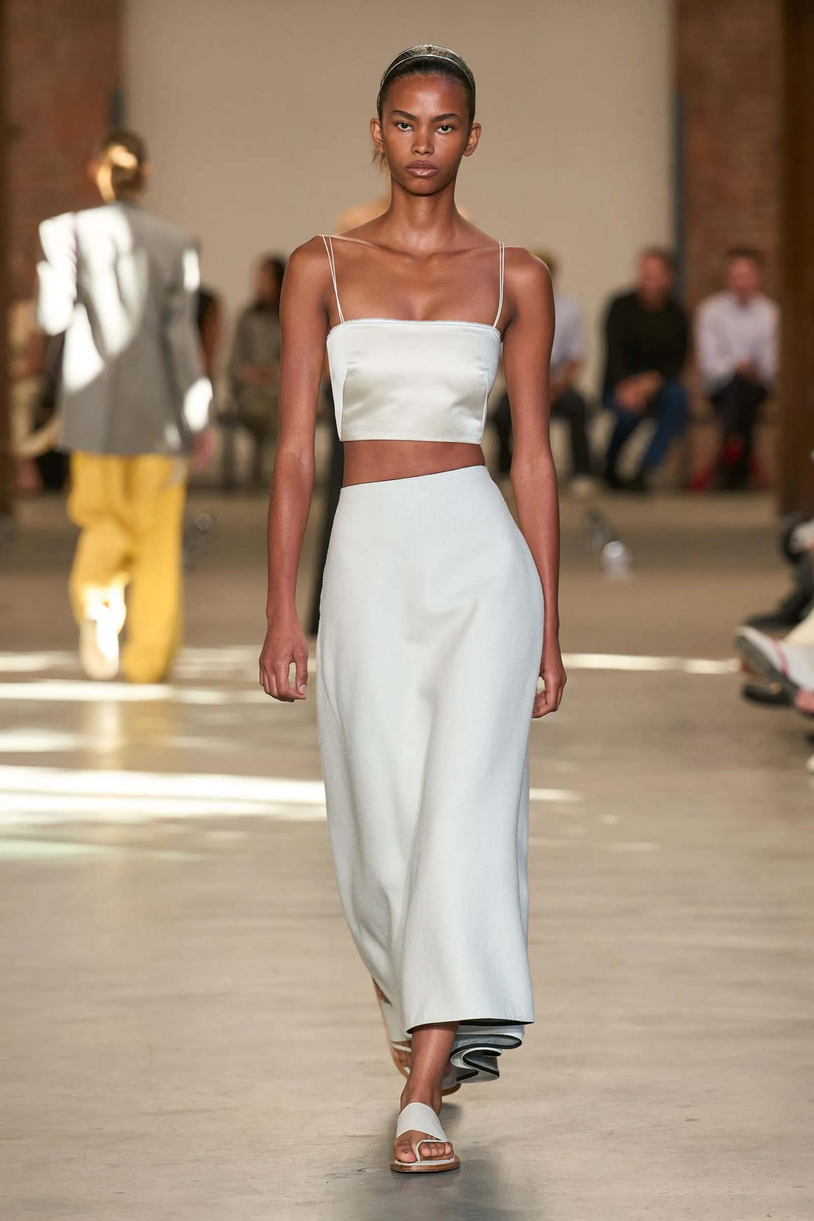 Model walks a runway in a pale blue crop top with thin straps and a matching high-waisted skirt with white sandals. Audience is blurred in the background.