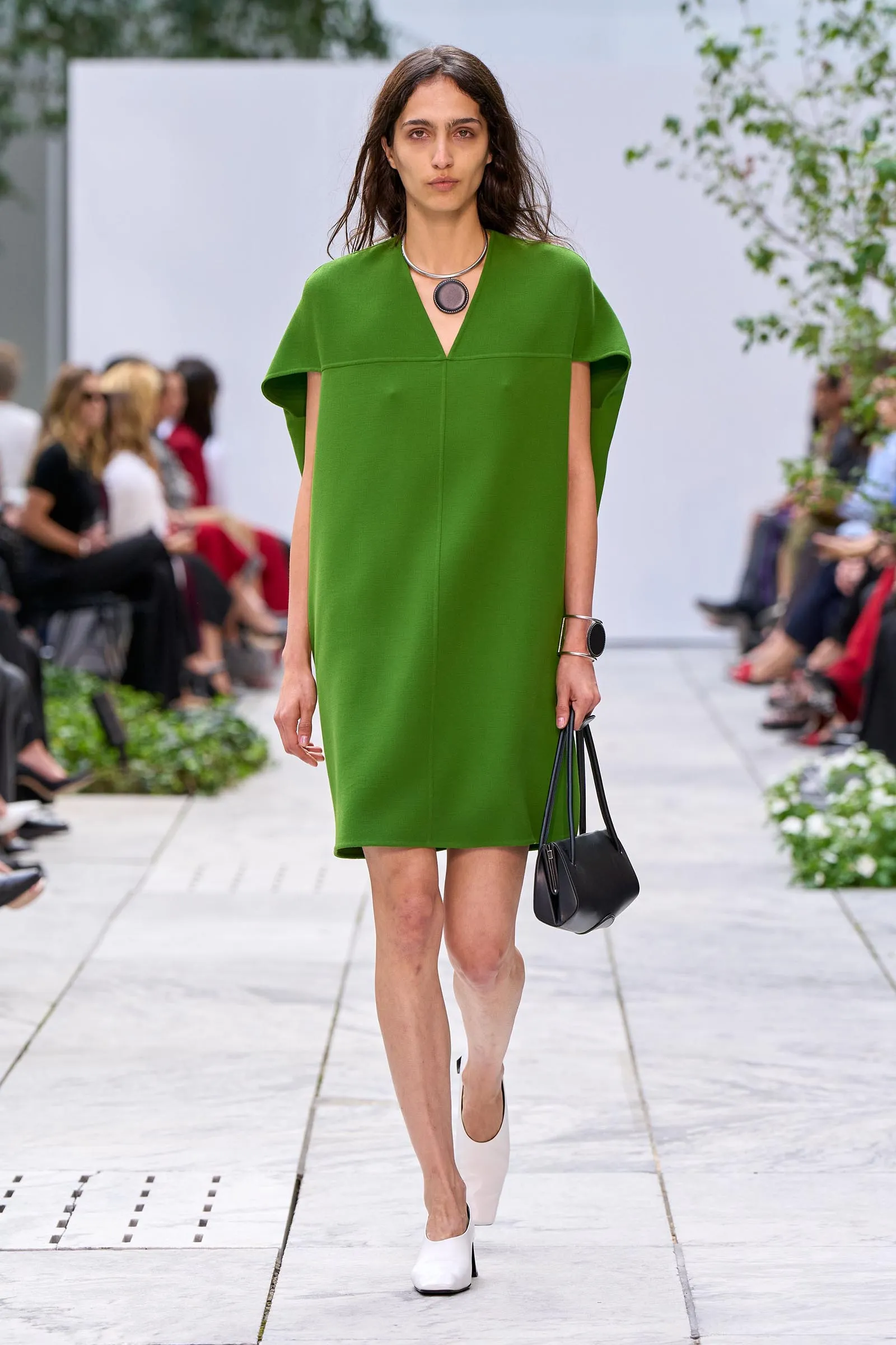 Model walking a runway in a bright green knee-length dress and white ankle boots, carrying a black handbag.