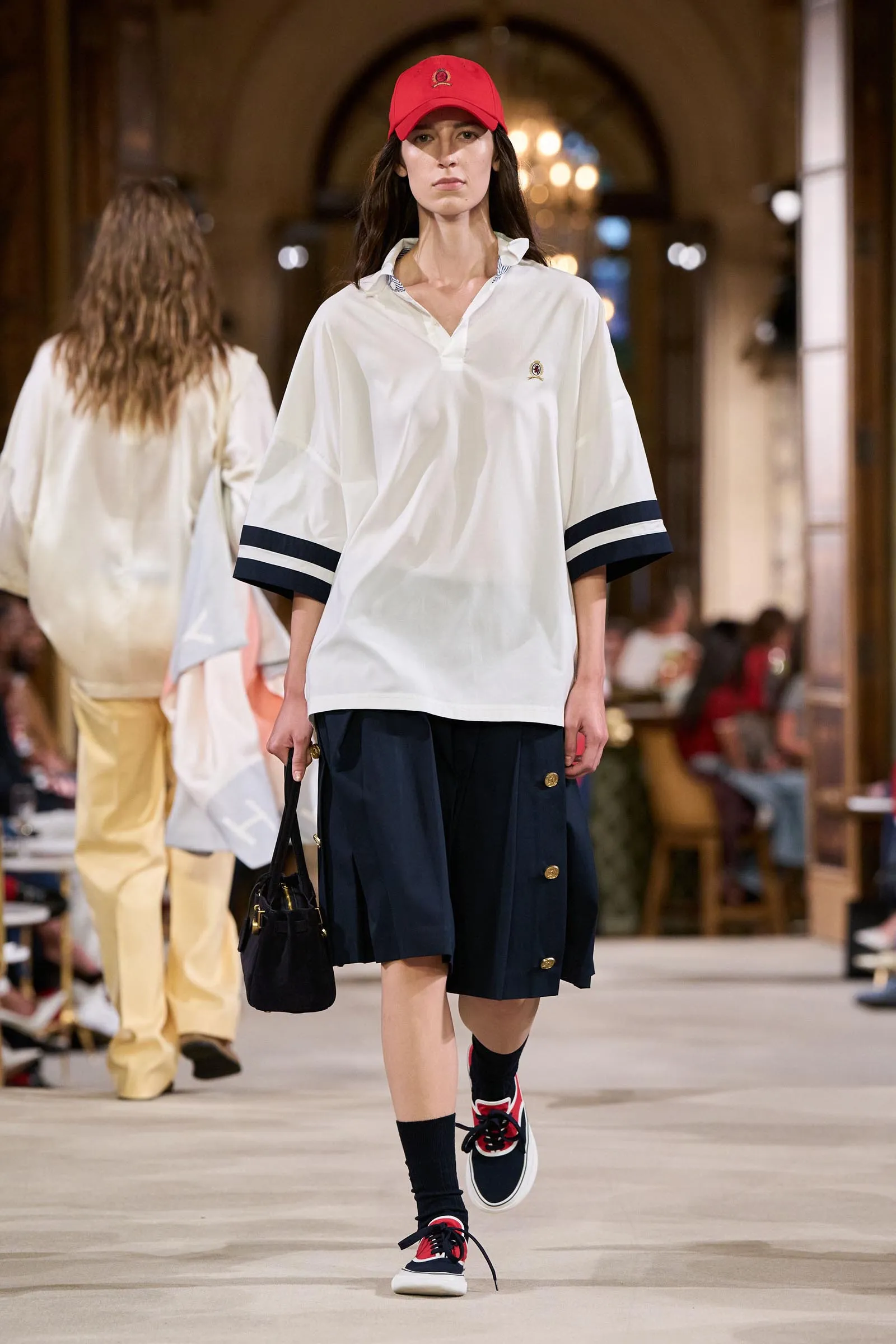 Model on a fashion runway wearing a loose white polo with navy sleeve stripes, navy shorts, red cap, and black-red sneakers.