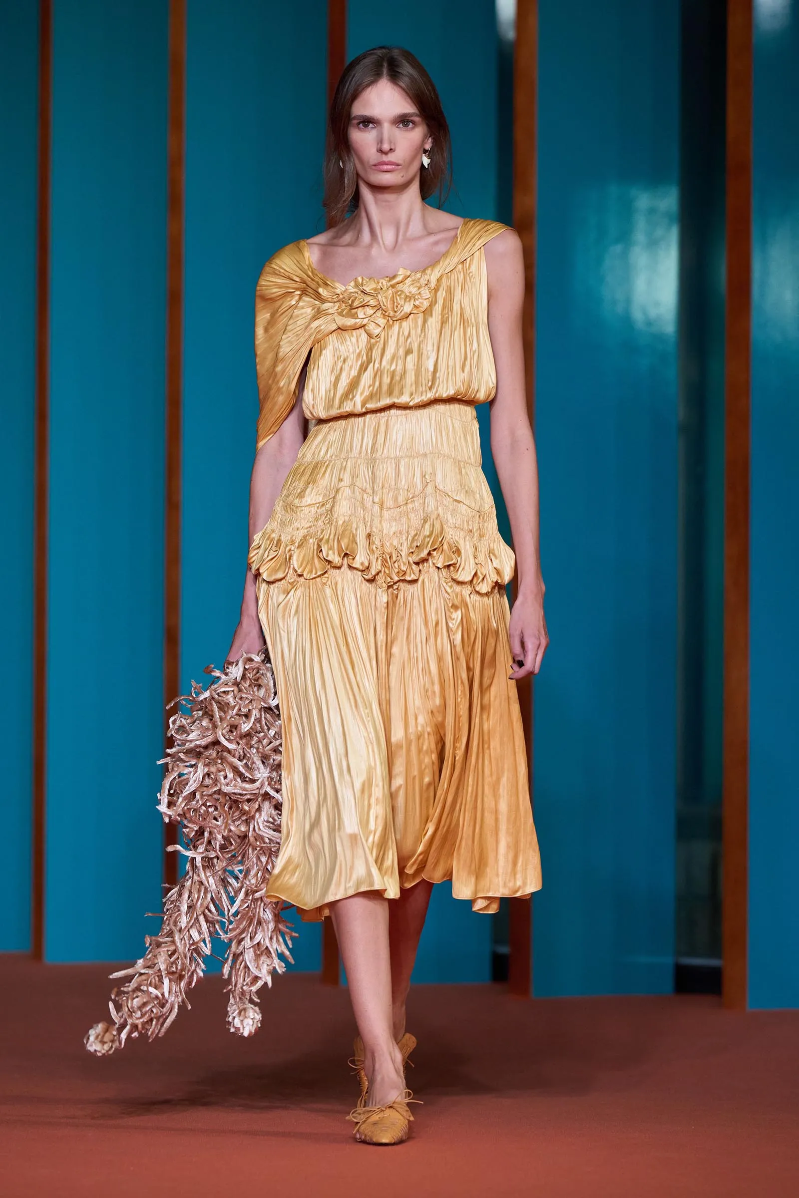 Model walking a fashion runway in a gold pleated dress, holding a textured beige bag against teal backdrop.